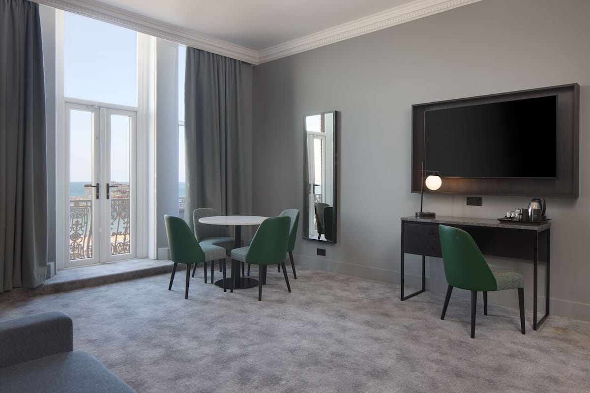 A modern hotel room with a balcony, a round dining table with chairs, a desk with a flat-screen TV, and a large mirror.