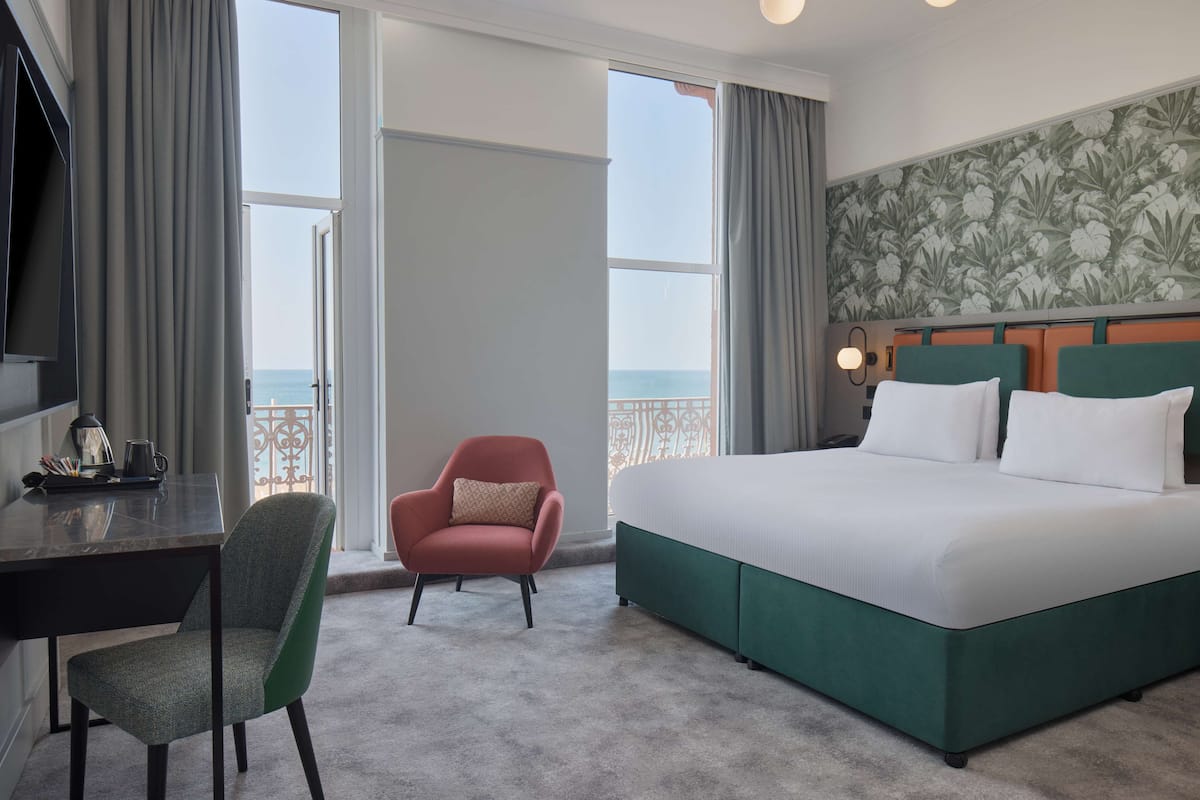 A hotel room with a large bed, a desk with a chair, a view of the sea through a balcony, and a floral wallpaper on the wall.