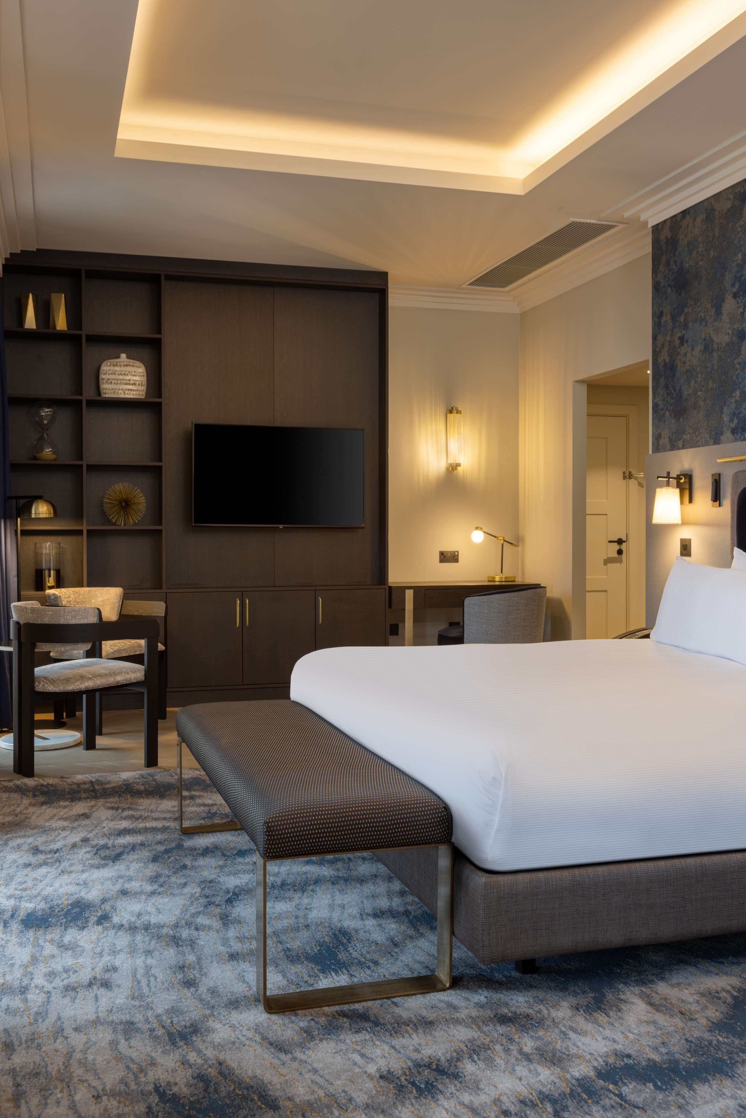 Una habitación de hotel moderna con una cama grande, un escritorio con una silla, un banco, un televisor de pantalla plana montado en la pared y una mesita de noche con una lámpara.