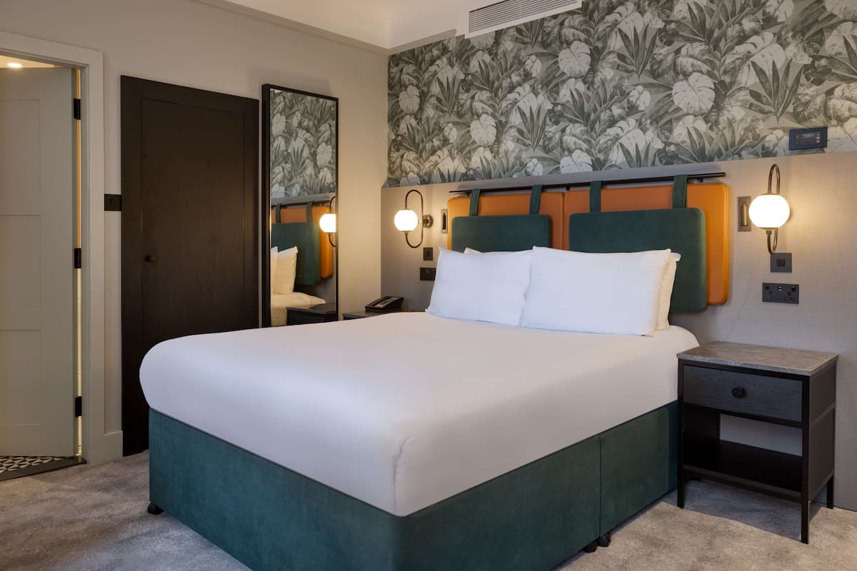 A hotel room with a large bed, a nightstand, a mirror, and a wall with floral wallpaper.