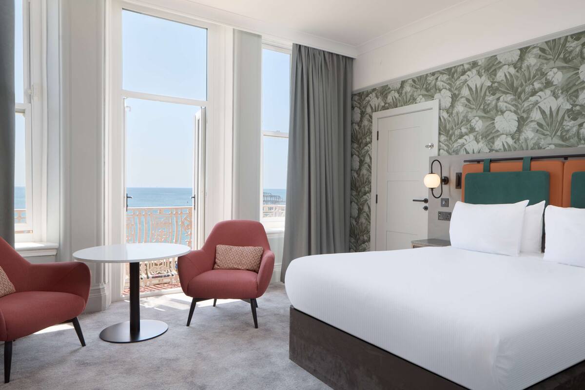 A hotel room with a large bed, two armchairs, a small round table, and a view of the sea.