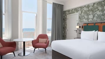 A hotel room with a large bed, two armchairs, a small round table, and a view of the sea.