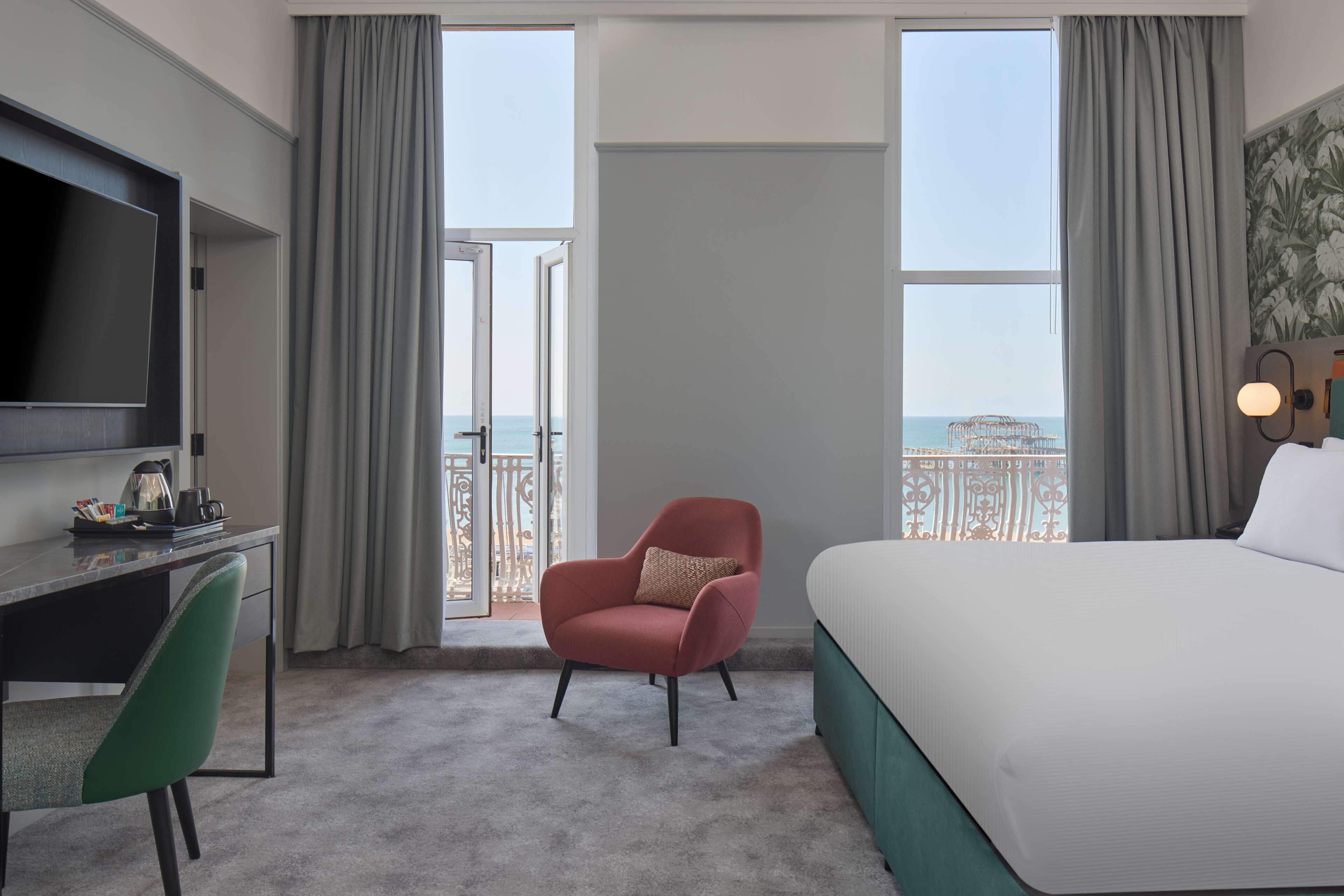 A hotel room with a bed, a desk with a TV, a chair, and a view of the sea through large windows.