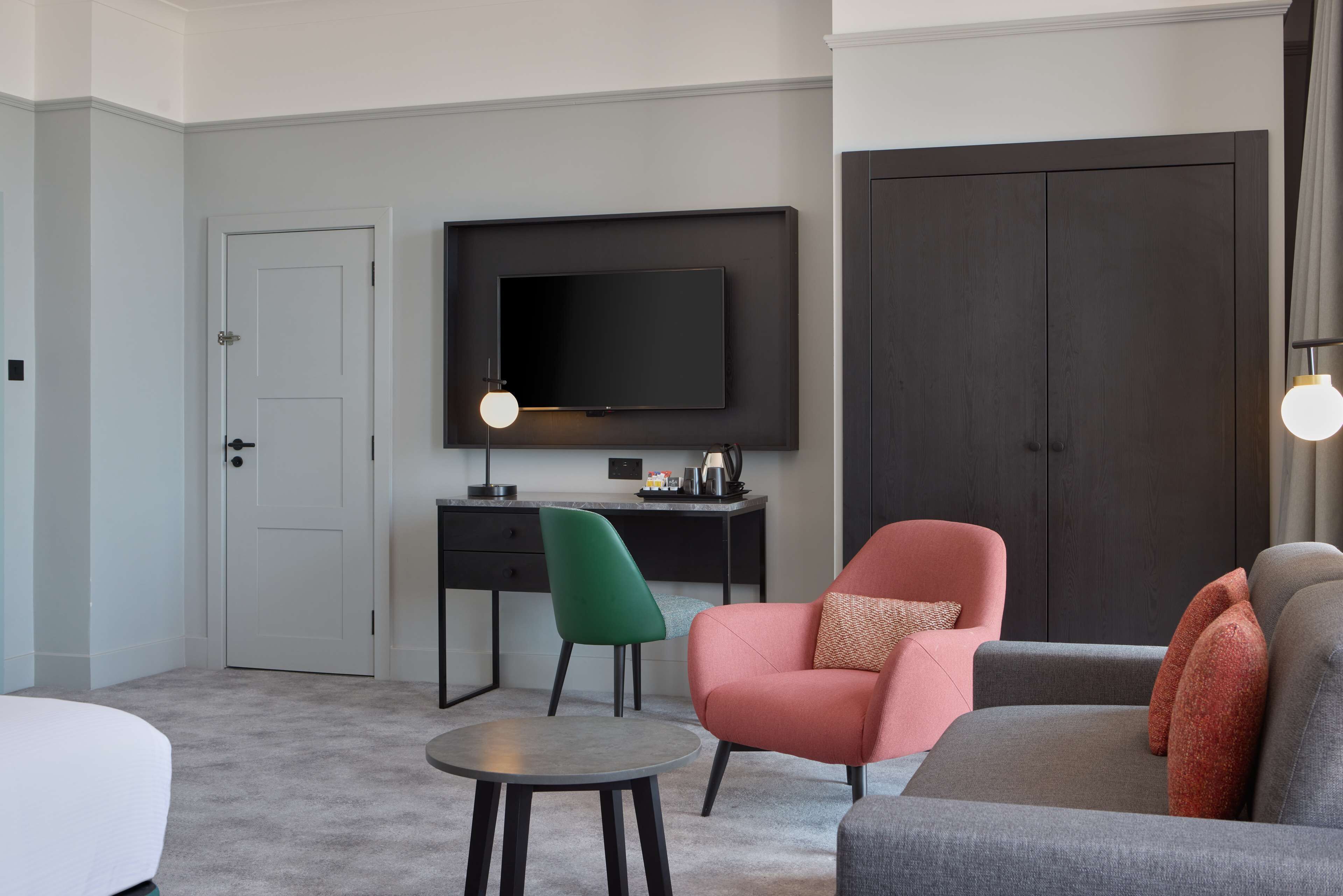 Ein modernes Wohnzimmer mit einem an der Wand befestigten Flachbildfernseher, einem grauen Sofa, einem pinken Sessel, einem grünen Stuhl und einem kleinen runden Tisch.
