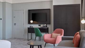 A modern living room with a flat-screen TV mounted on the wall, a grey sofa, a pink armchair, a green chair, and a small round table.