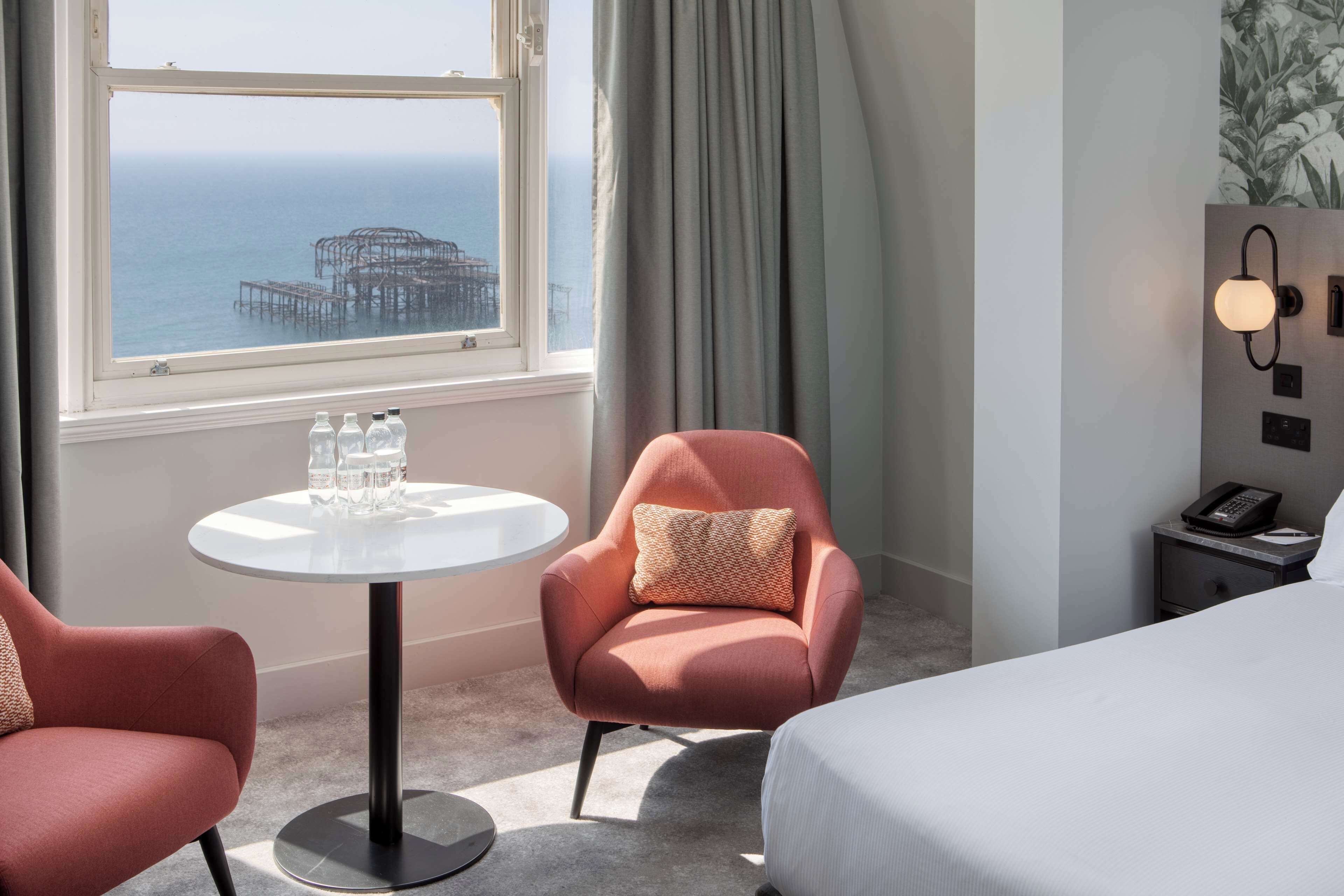 A hotel room with a bed, a round table with two chairs, a view of the ocean, and a nightstand with a phone.