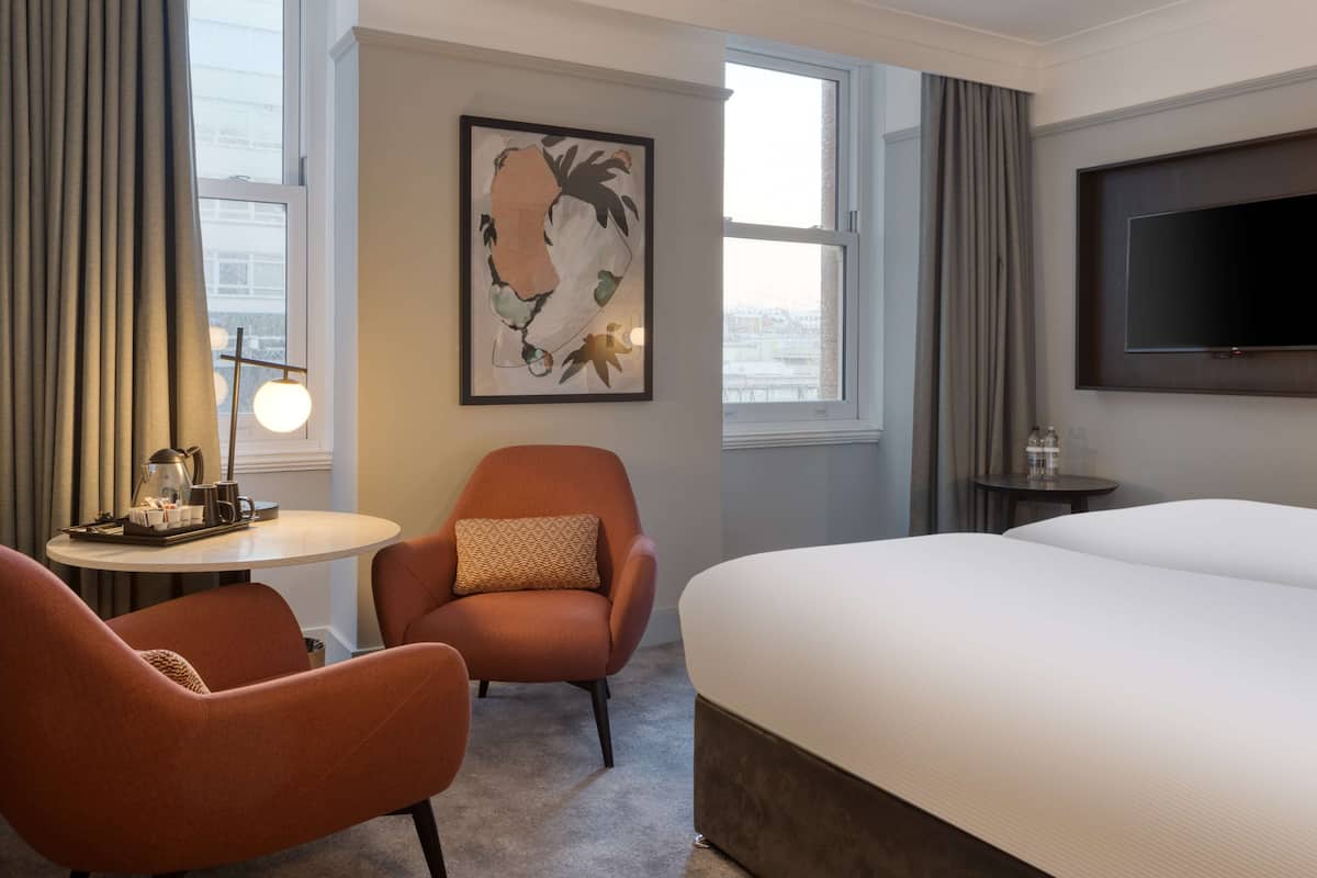 A hotel room with a large bed, two armchairs, a small round table with a lamp, a painting on the wall, and a TV mounted on the wall.