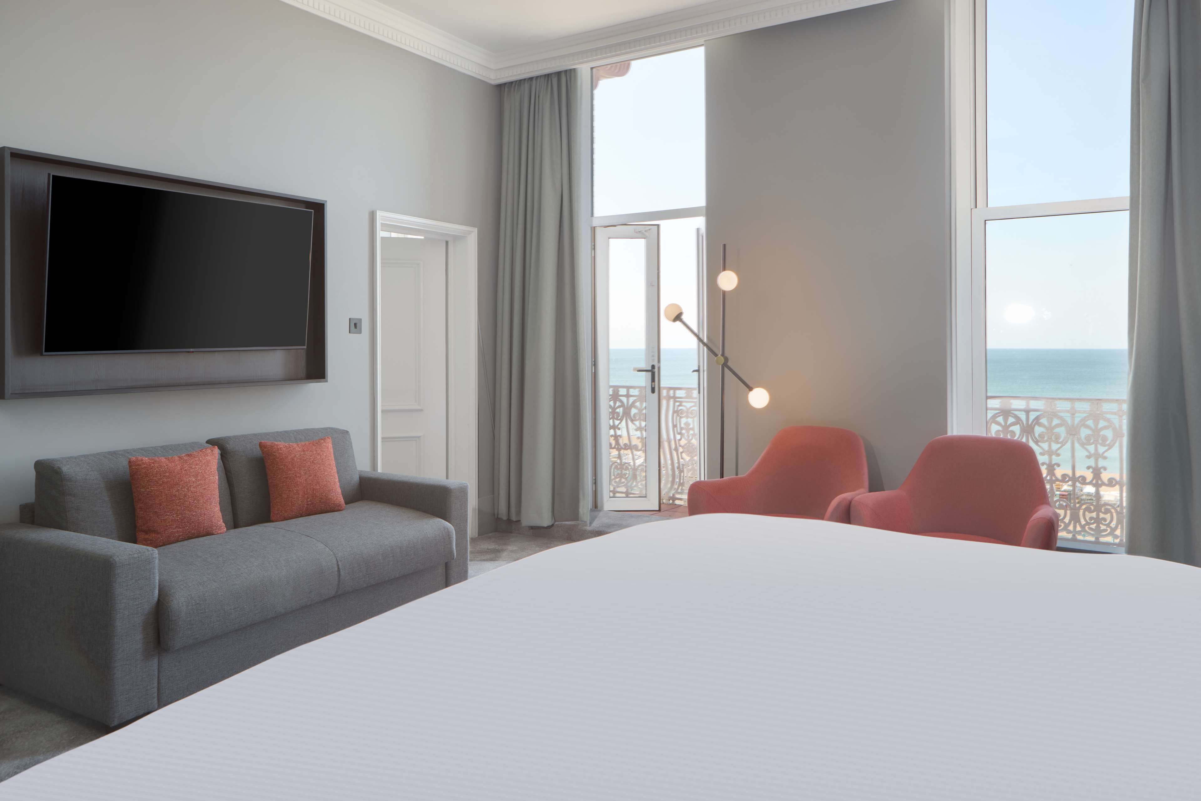 Een hotelkamer met een groot bed, een grijze bank met rode kussens, een flatscreen televisie en een balkon met uitzicht op zee.