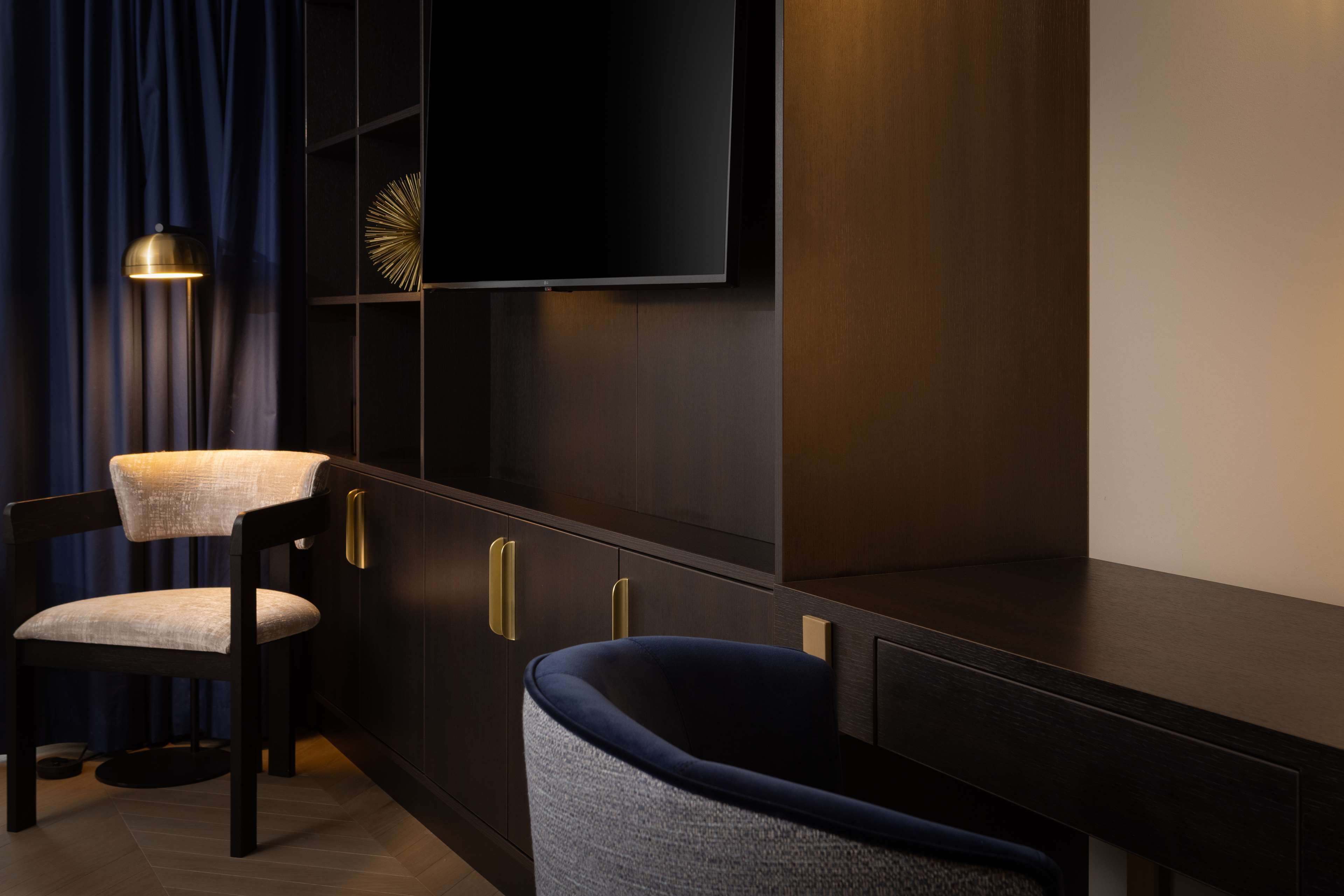Una habitación moderna con un armario de madera oscura, un televisor de pantalla plana en la pared, un sillón tapizado en azul oscuro y una lámpara de pie.