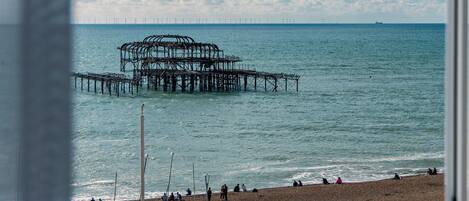 A coastal view with a pier extending into the sea, a beach with people and umbrellas, and a clear sky with clouds.