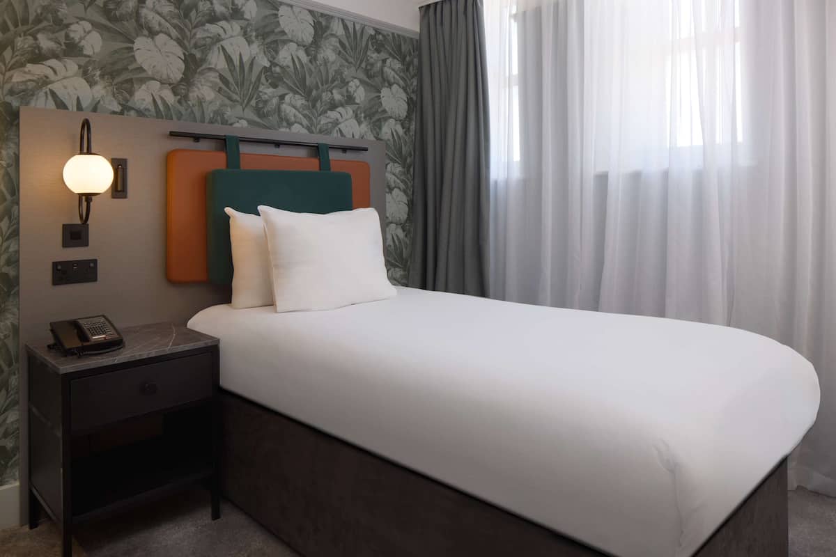 A hotel room with a bed, a nightstand with a phone, a wall-mounted lamp, and a window with curtains.