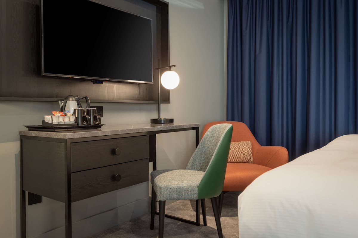 A hotel room with a bed, a desk with a lamp, a chair, and a television on the wall.
