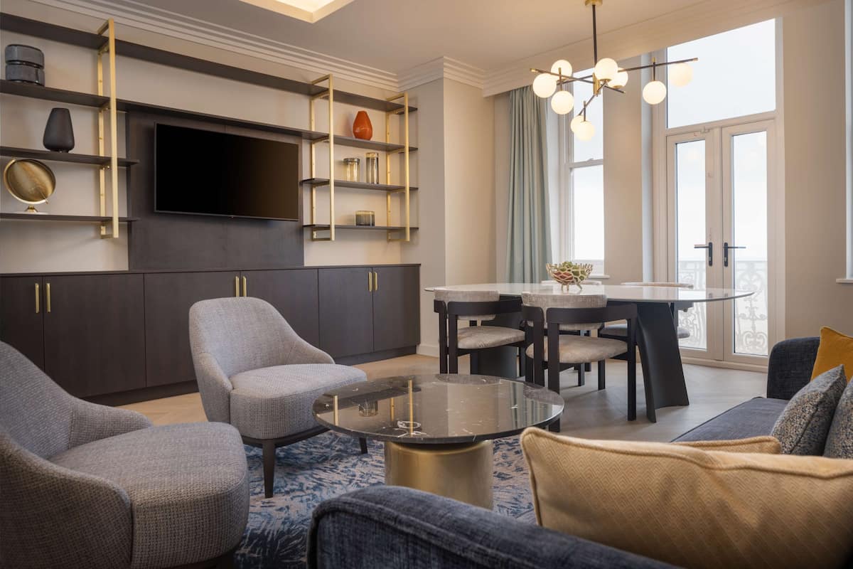 A modern living room with a flat-screen TV, dark cabinets, a round glass coffee table, and a dining area with a glass table and chairs.