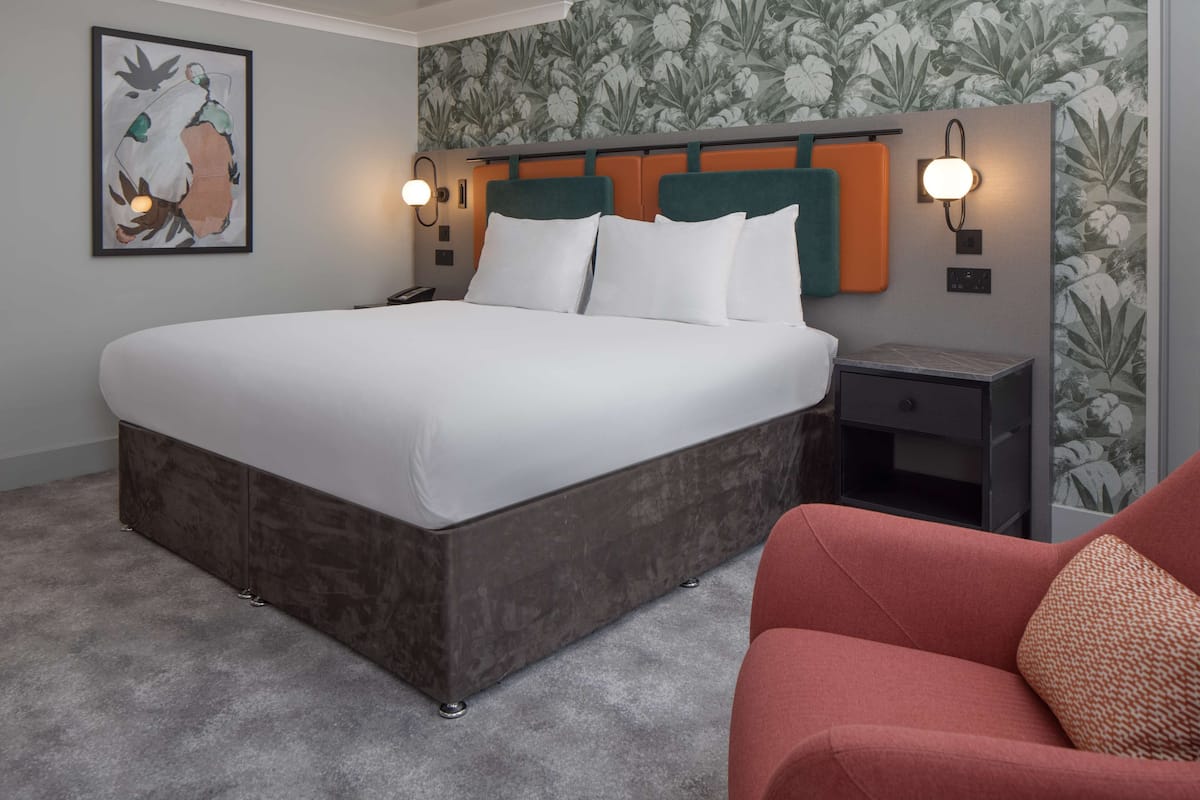 A hotel room with a large bed, a nightstand with a lamp, a red chair, and a wall with a floral wallpaper.