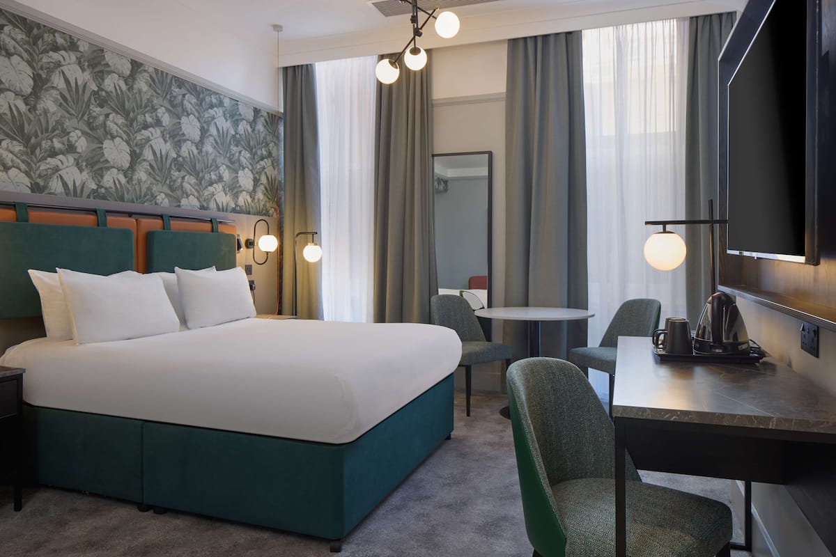 A hotel room with a large bed, a desk with a chair, a small table, and a television.