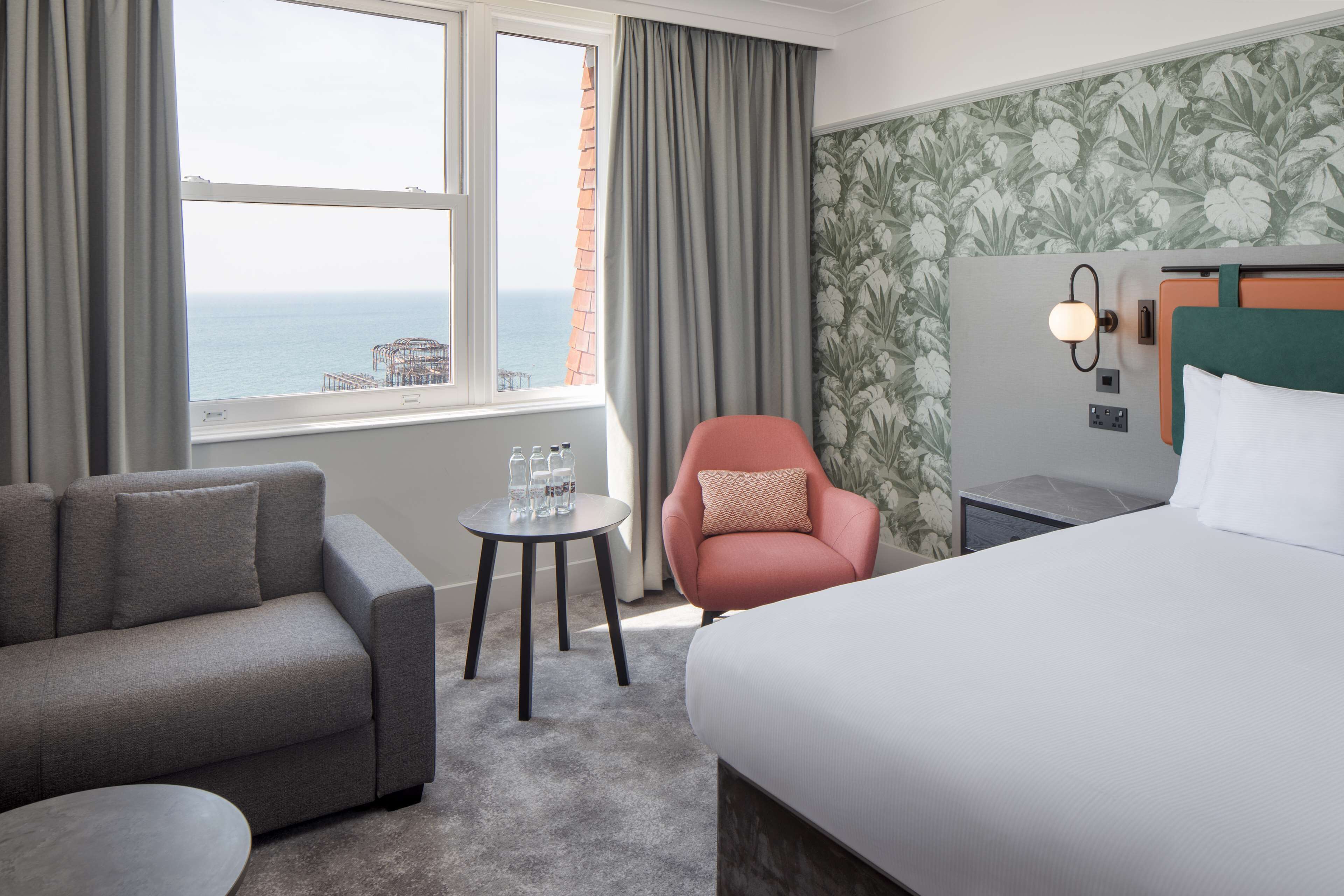 Ein Hotelzimmer mit grauem Sofa, einem rosa Sessel, einem kleinen runden Tisch mit Gläsern, einem Bett und Meerblick durch ein Fenster.