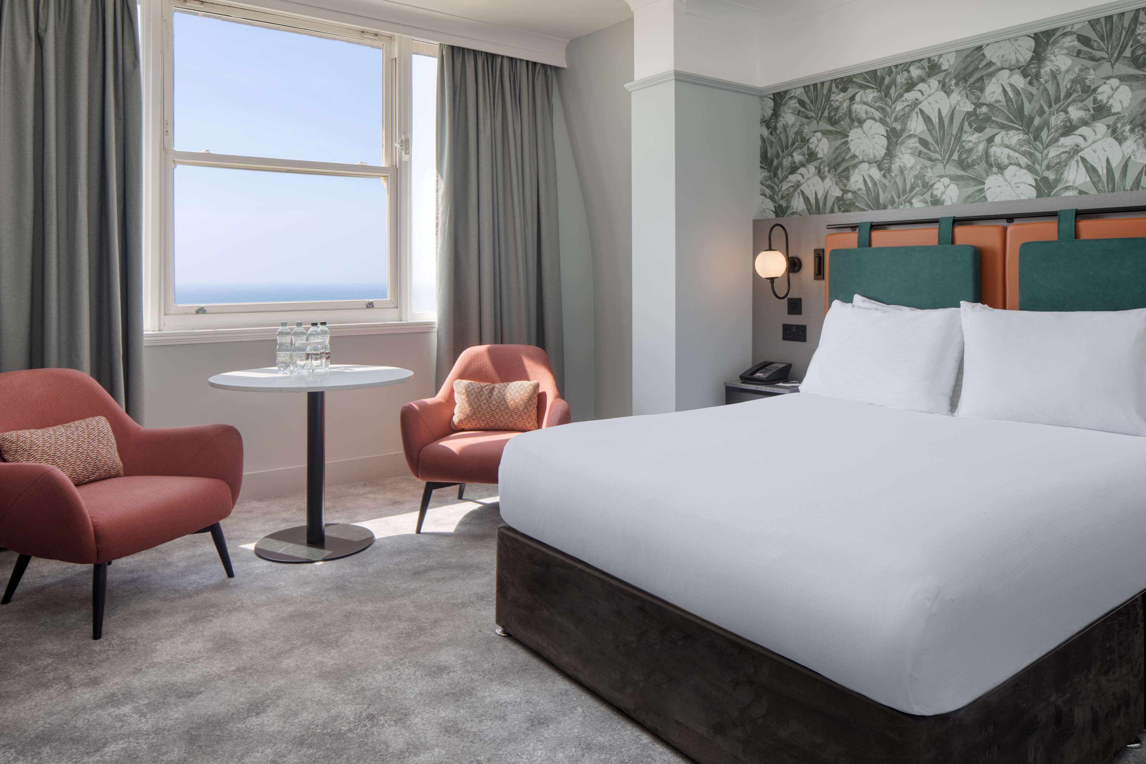 A hotel room with a large bed, two armchairs, a small round table with water bottles, a nightstand, and a view of the ocean through a window with curtains.
