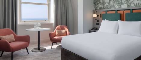 A hotel room with a large bed, two armchairs, a small round table with water bottles, a nightstand, and a view of the ocean through a window with curtains.