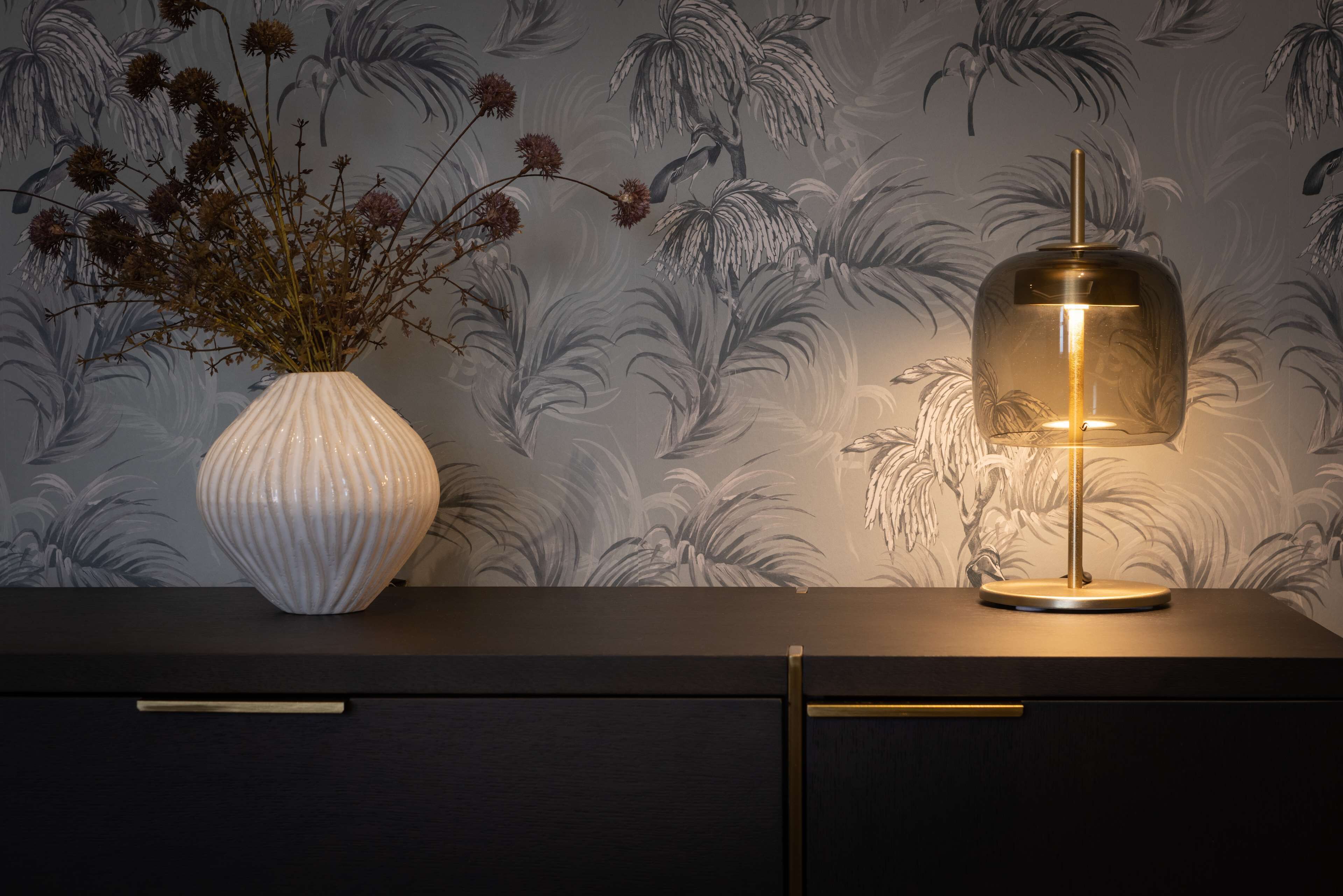 A dark wooden desk with a white vase containing dried flowers and a modern lamp with a gold base and clear shade.