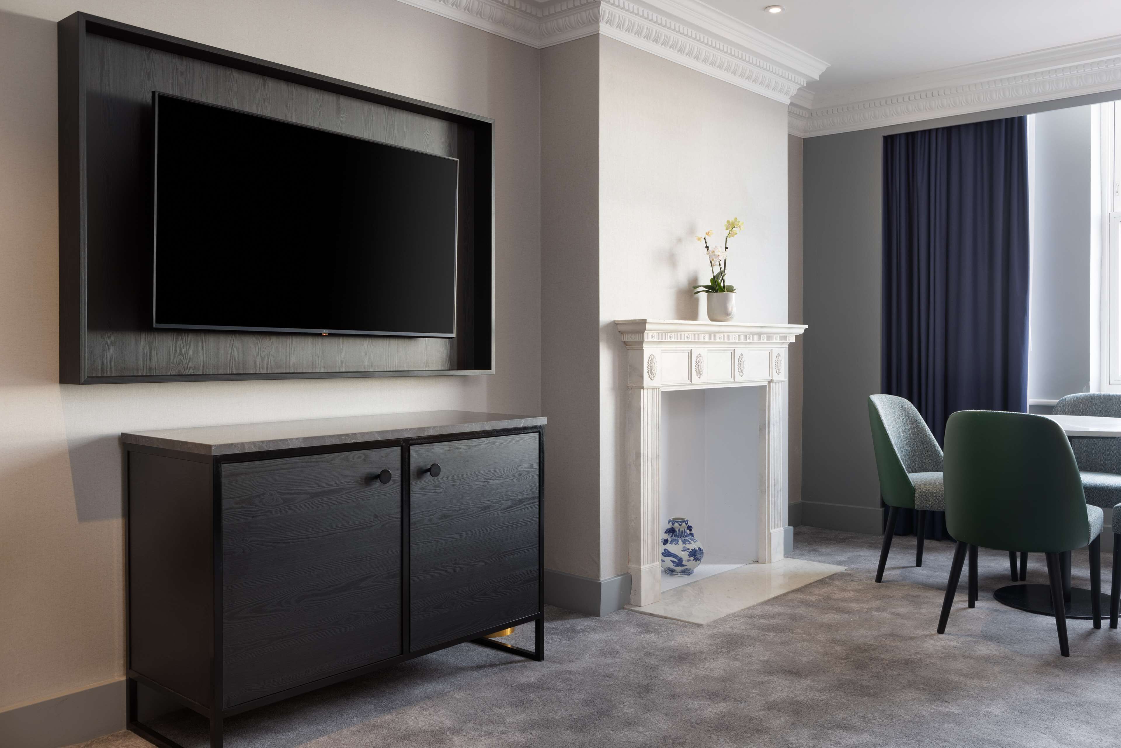 A modern living room with a flat-screen TV mounted on the wall, a dark wood cabinet, a white fireplace, and two green chairs.