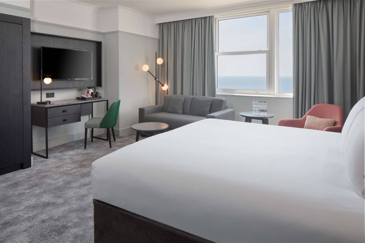 A hotel room with a bed, a desk with a chair, a sofa, a TV, and a window with curtains.