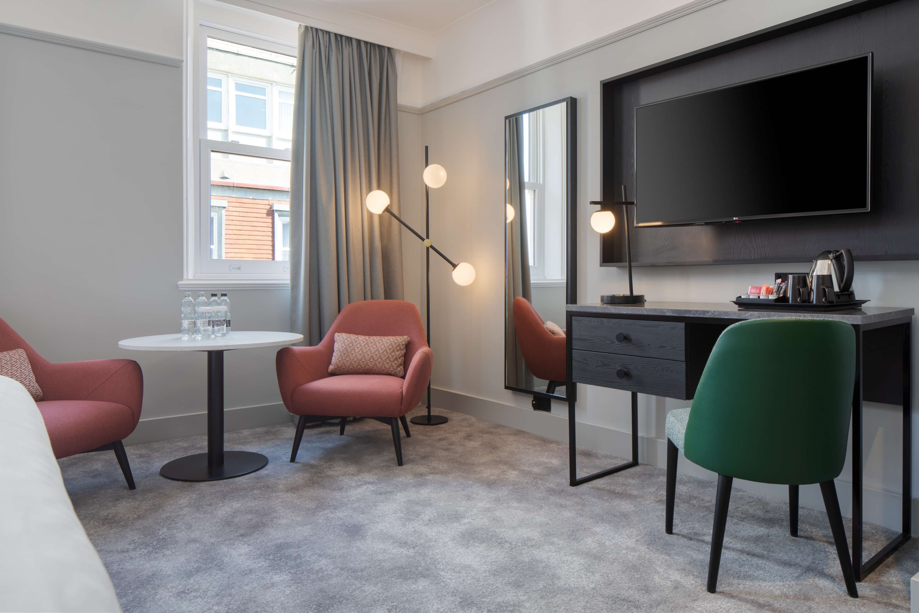 A hotel room with a bed, a desk, a chair, a round table, a TV, and a large window with curtains.