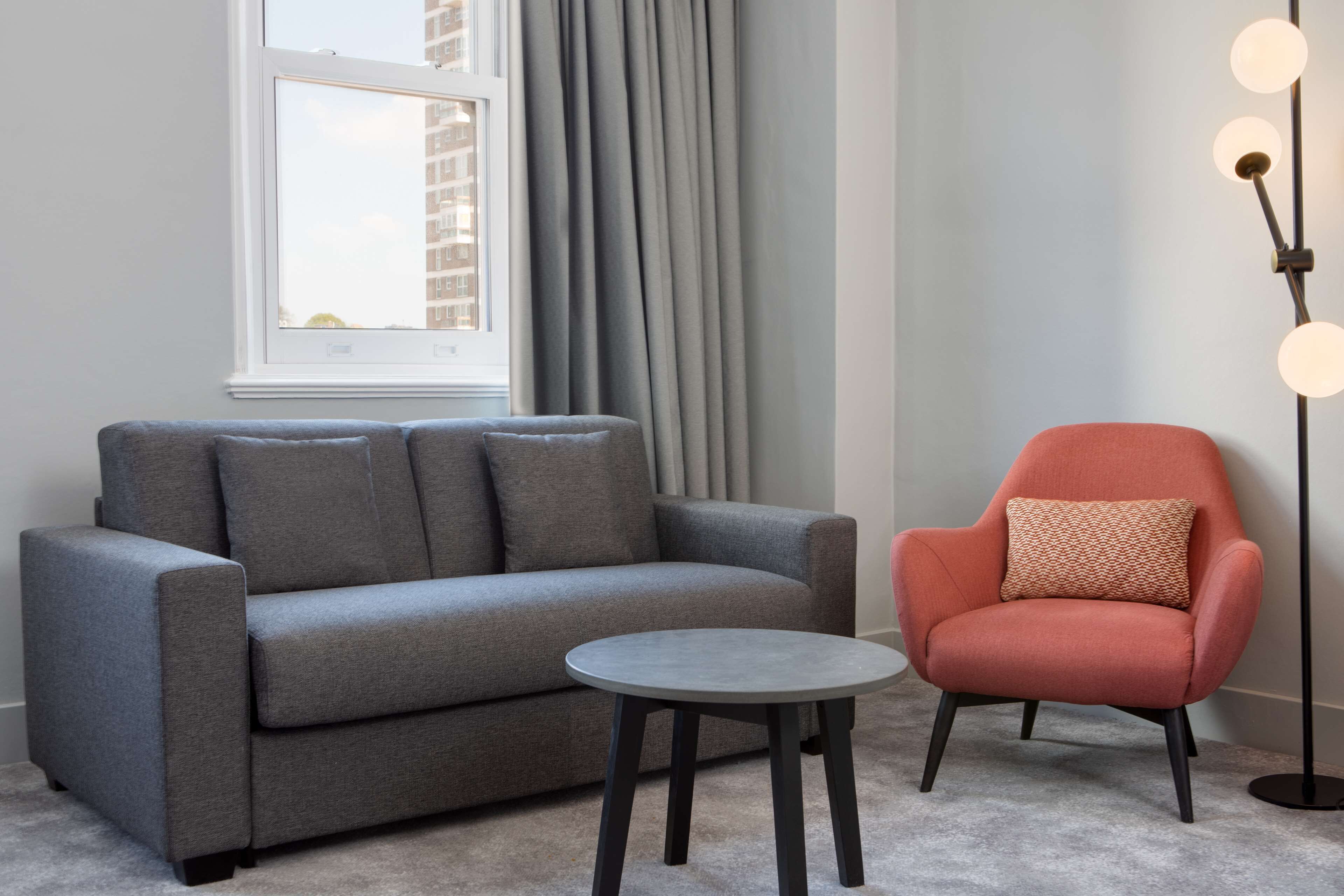 A grey sofa, a small round table, and a red armchair in a room with a window and a floor lamp.