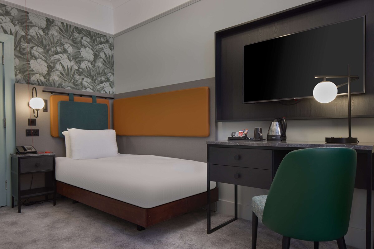 A modern bedroom with a bed, bedside table, lamp, and a desk with a chair.