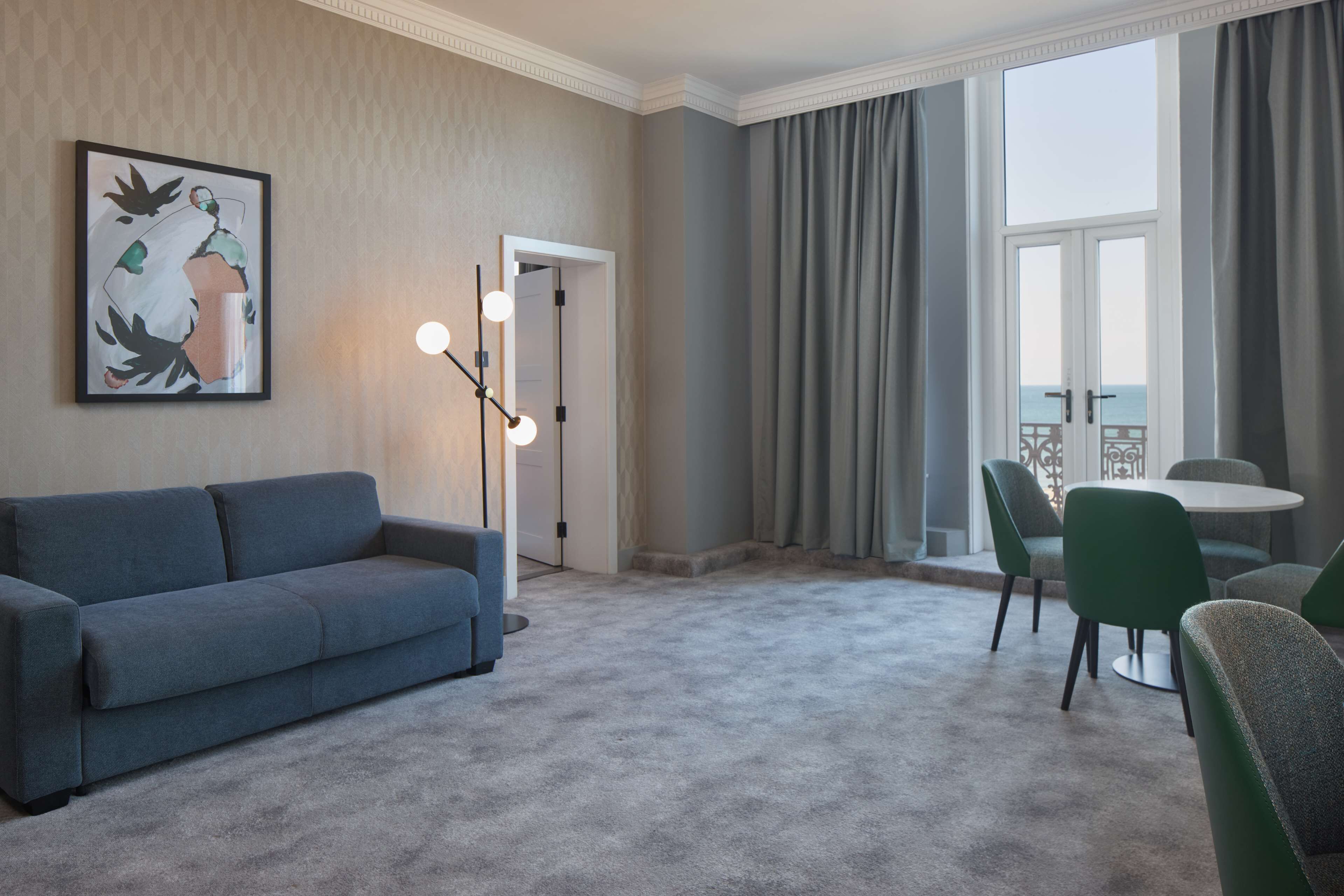 Et hotelværelse med gråt tæppe, en blå sofa, et spisebord med grønne stole, et stort vindue med gardiner og et indrammet kunstværk på væggen.