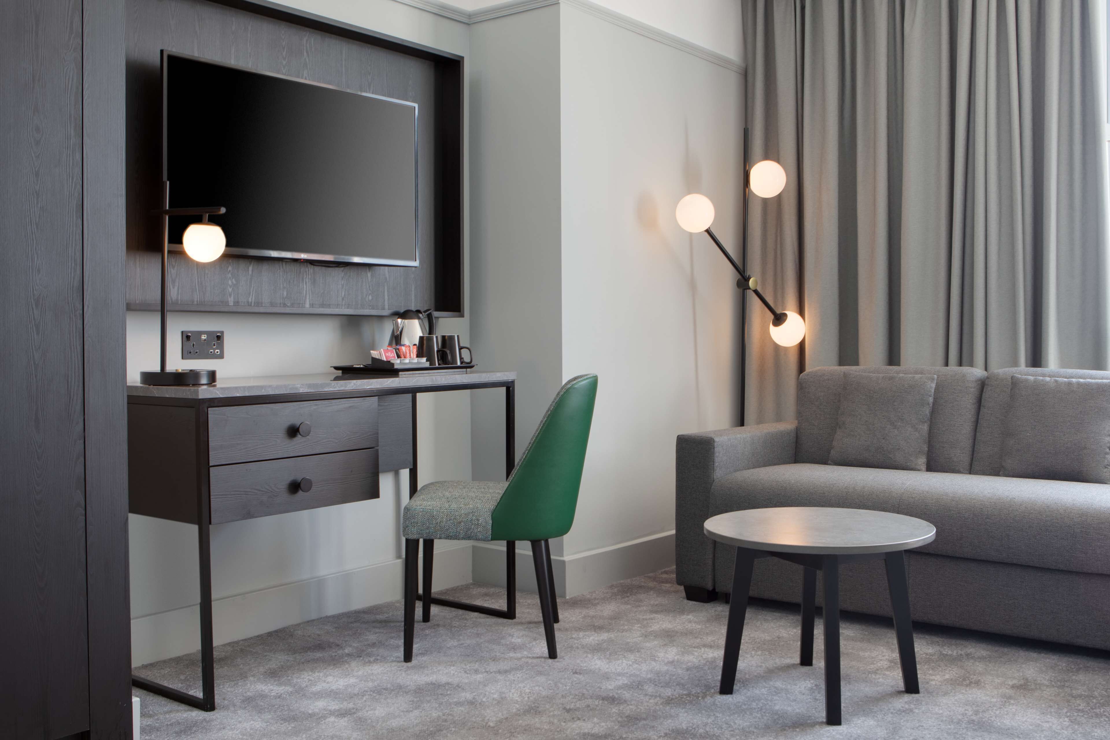 A modern hotel room with a dark wood desk, a green upholstered chair, a grey sofa, and a round coffee table.