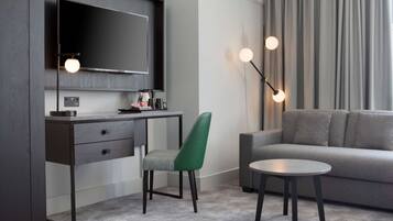A modern hotel room with a dark wood desk, a green upholstered chair, a grey sofa, and a round coffee table.