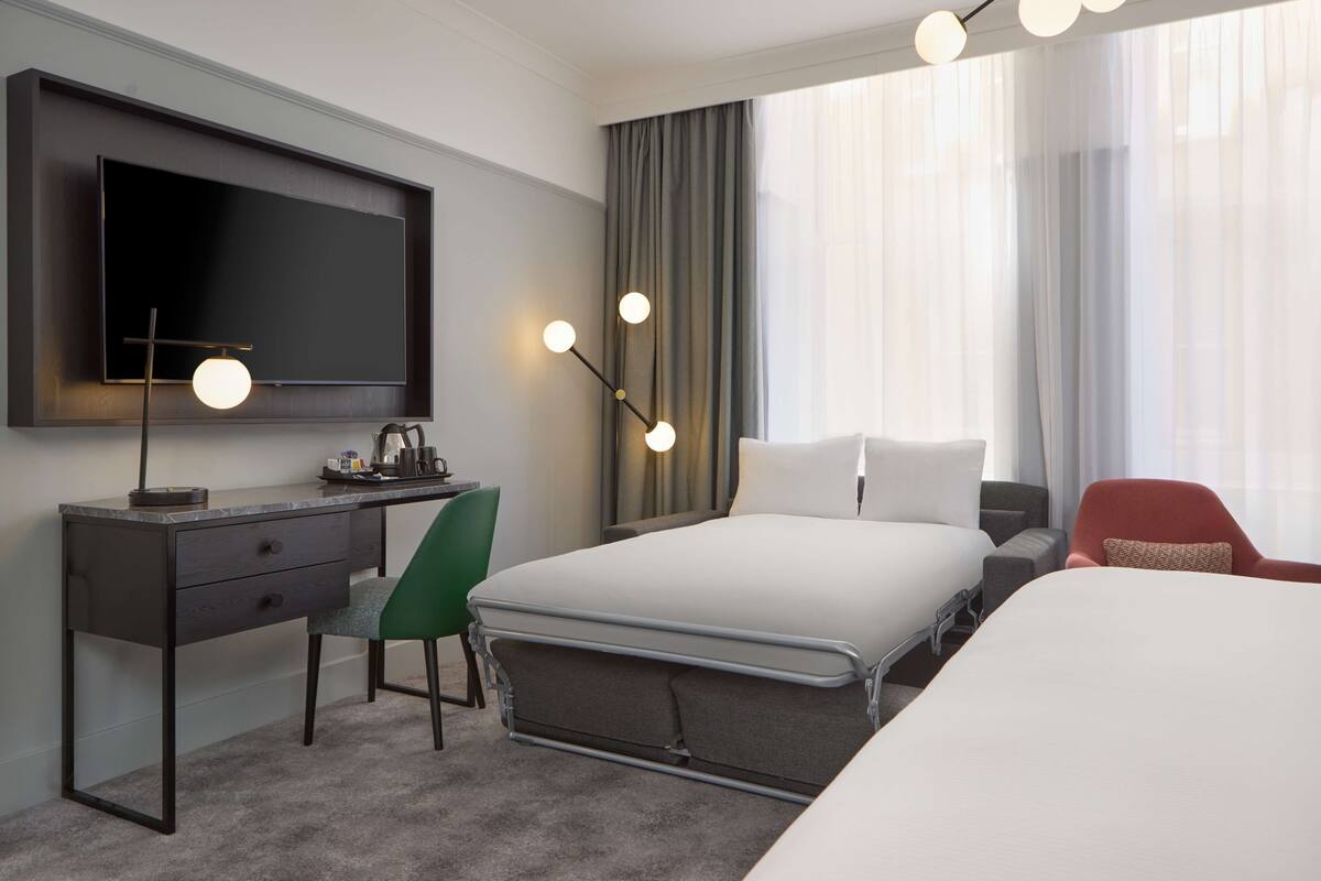 A hotel room with a large bed, a desk with a flat-screen TV, a green chair, and a red armchair.