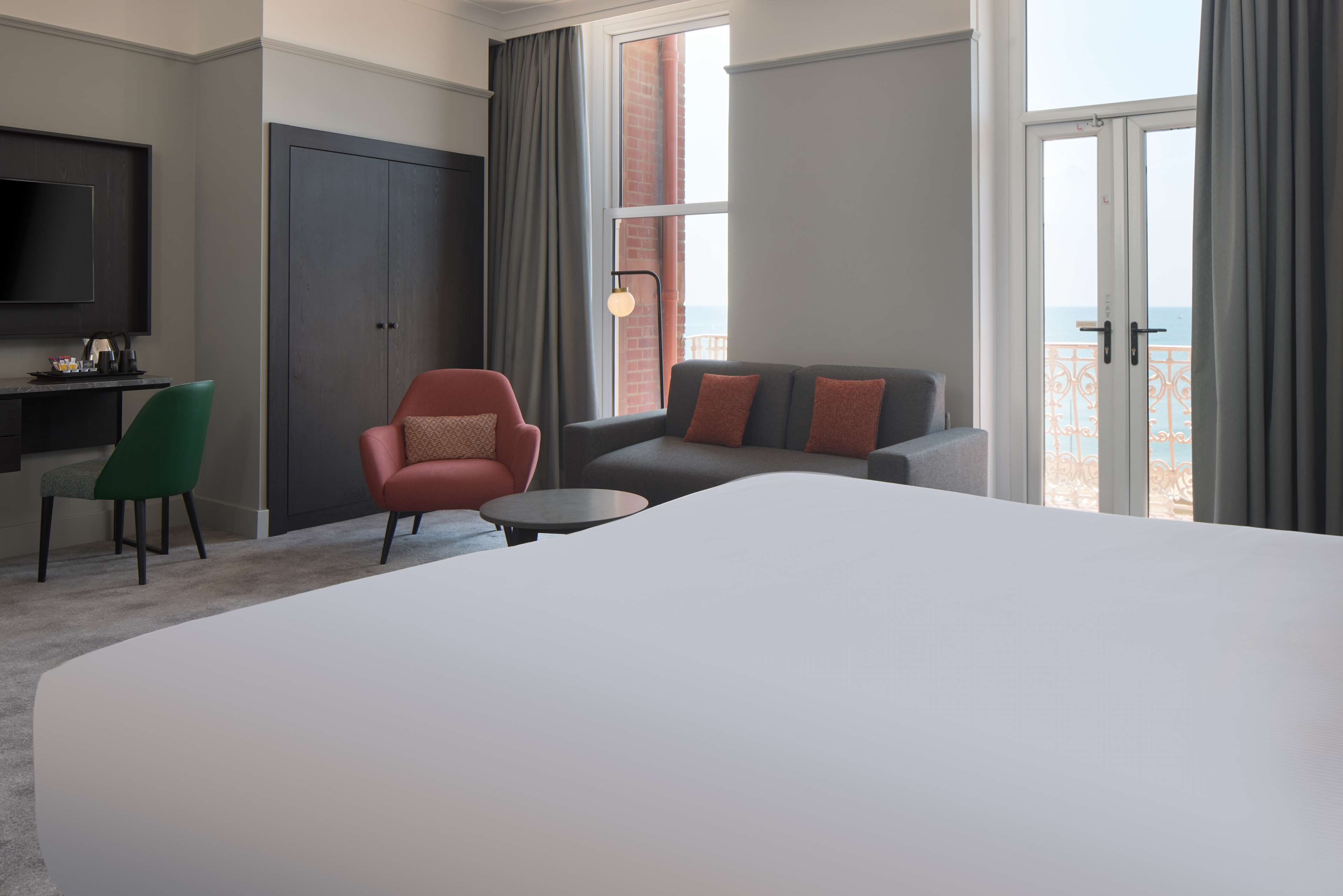 A hotel room with a large bed, a sofa, a chair, a small table, a TV, and a window with a view of the sea.