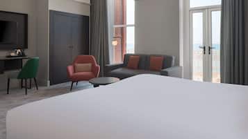 A hotel room with a large bed, a sofa, a chair, a small table, a TV, and a window with a view of the sea.