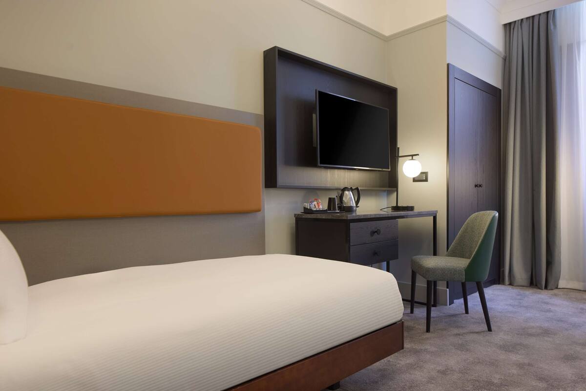 A hotel room with a bed, a desk with a flat-screen TV, a chair, and a wardrobe.