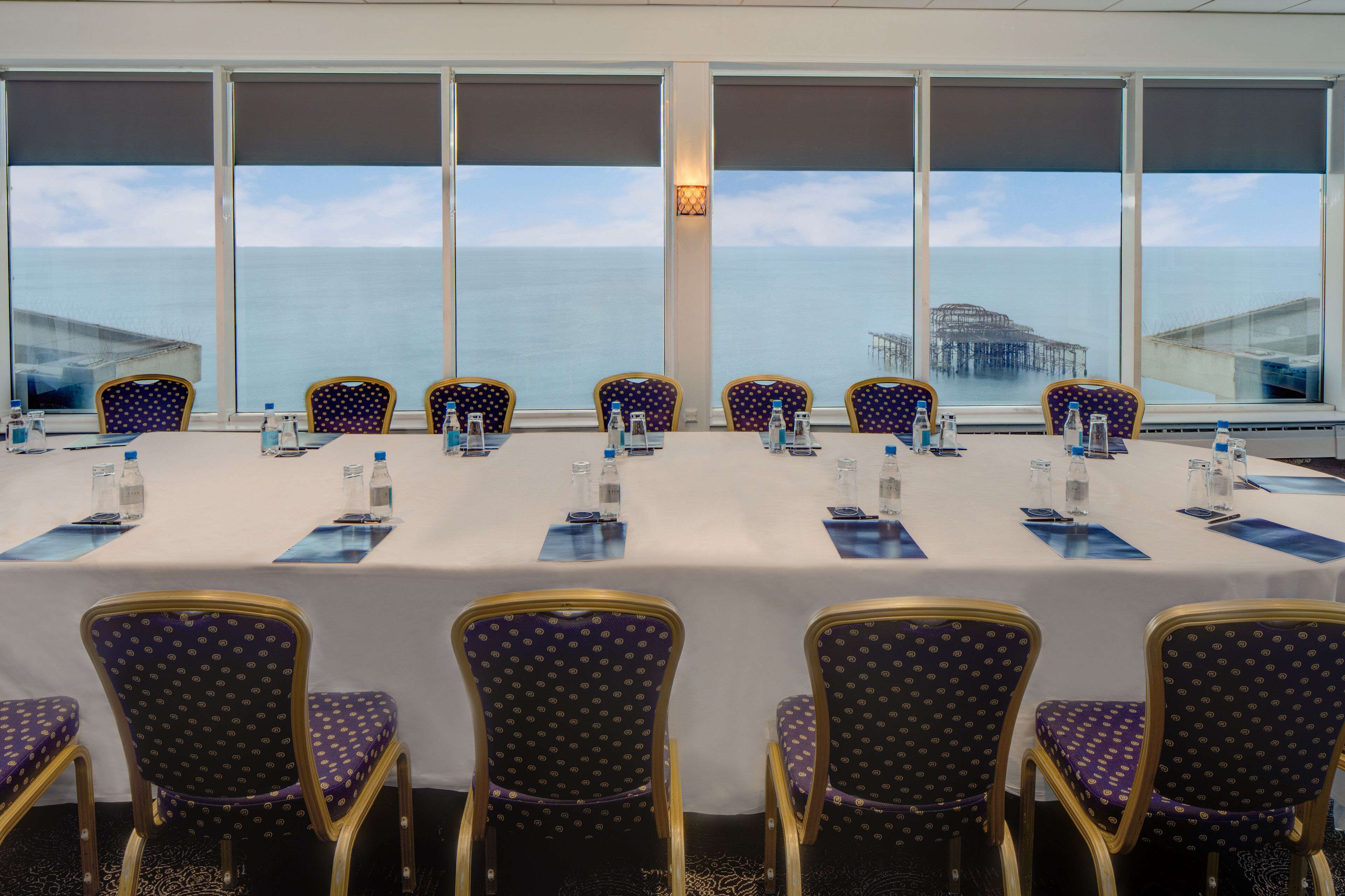 Un salón de conferencias con vista al océano y un muelle, equipado con mesas, sillas y botellas de agua.