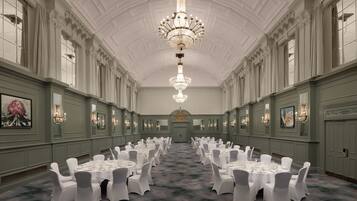 A formal banquet hall with rows of round tables, white chairs, and chandeliers.