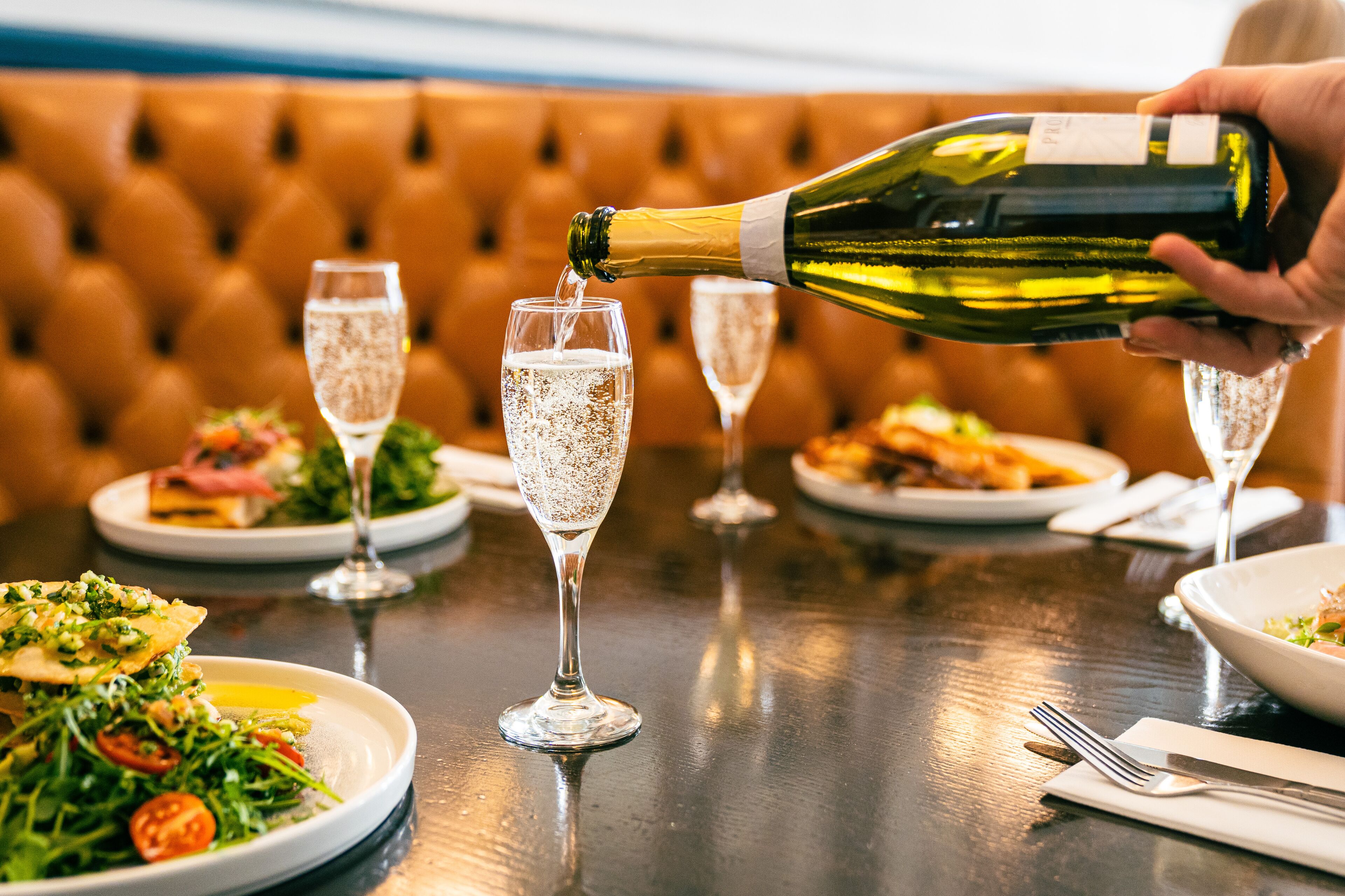 A table with multiple glasses of champagne, a bottle being poured, and plates of food.
