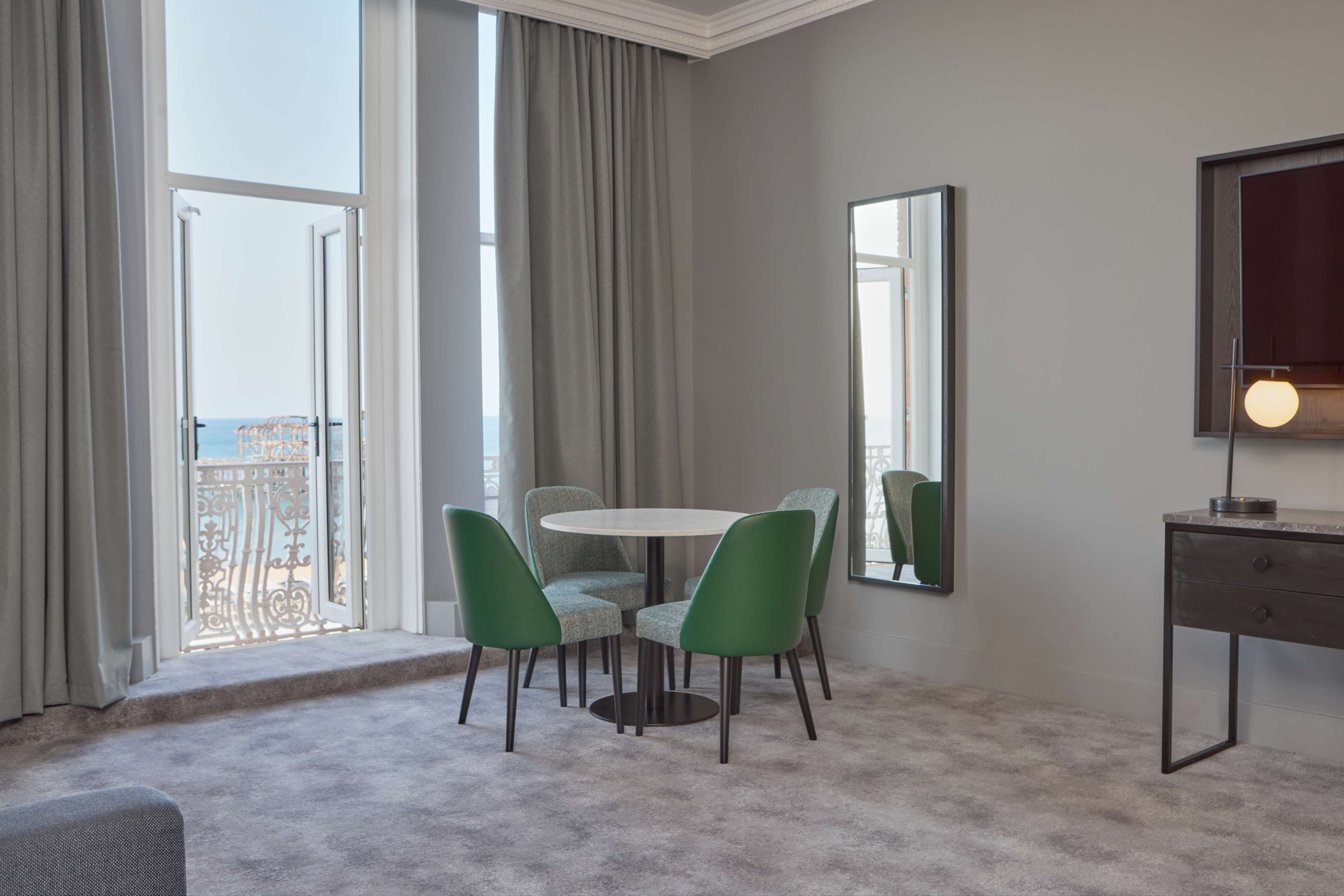 A room with a round table and four chairs, a large mirror, a TV, and a view of the sea through a balcony door.
