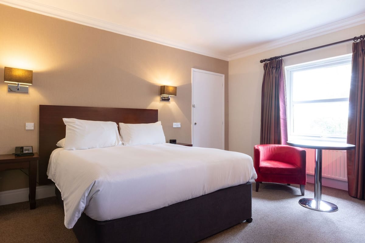 A hotel room with a large bed, two bedside lamps, a red chair, a small table, and a window with curtains.