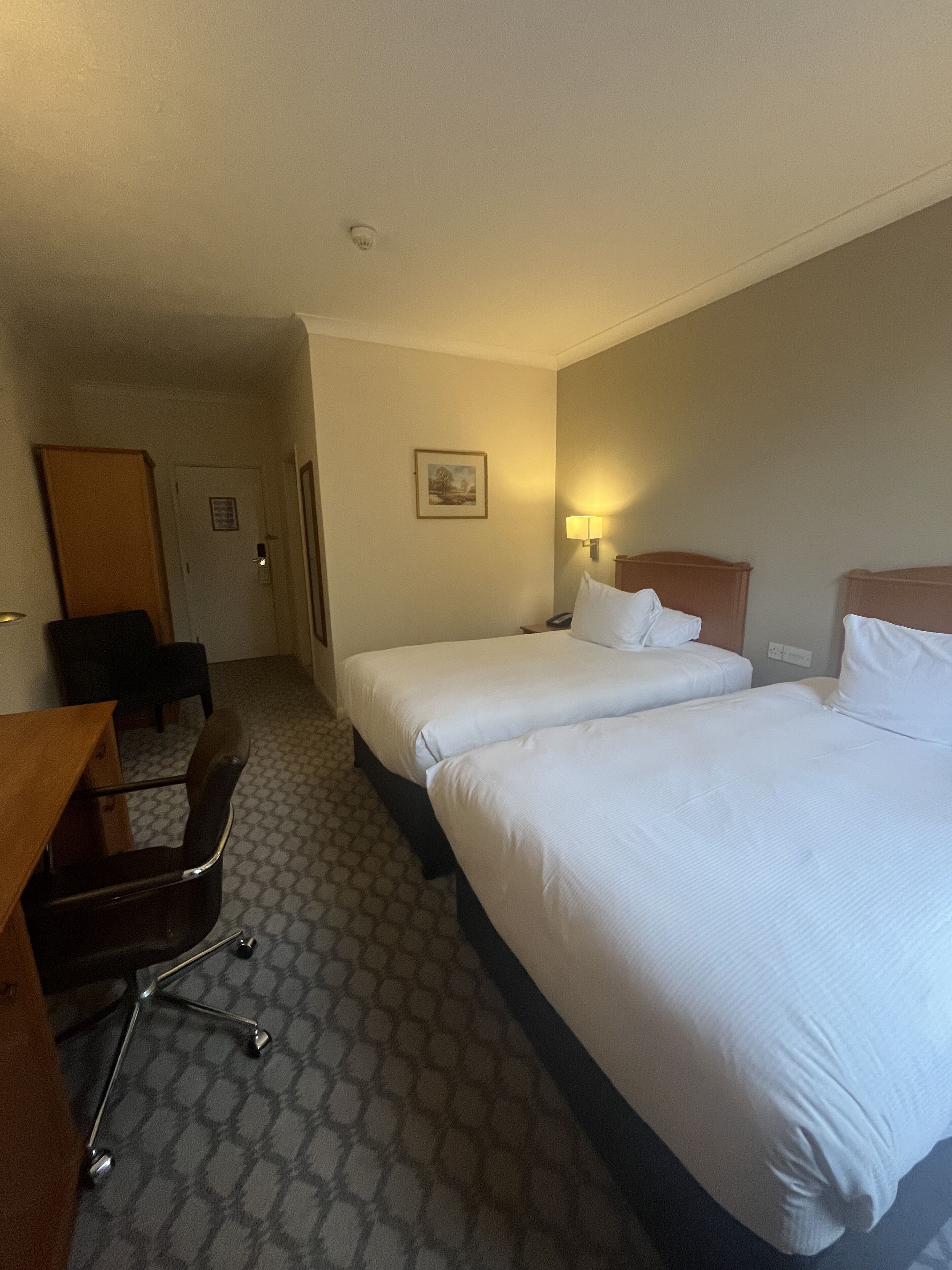 A hotel room with two beds, a desk, a chair, and a door.
