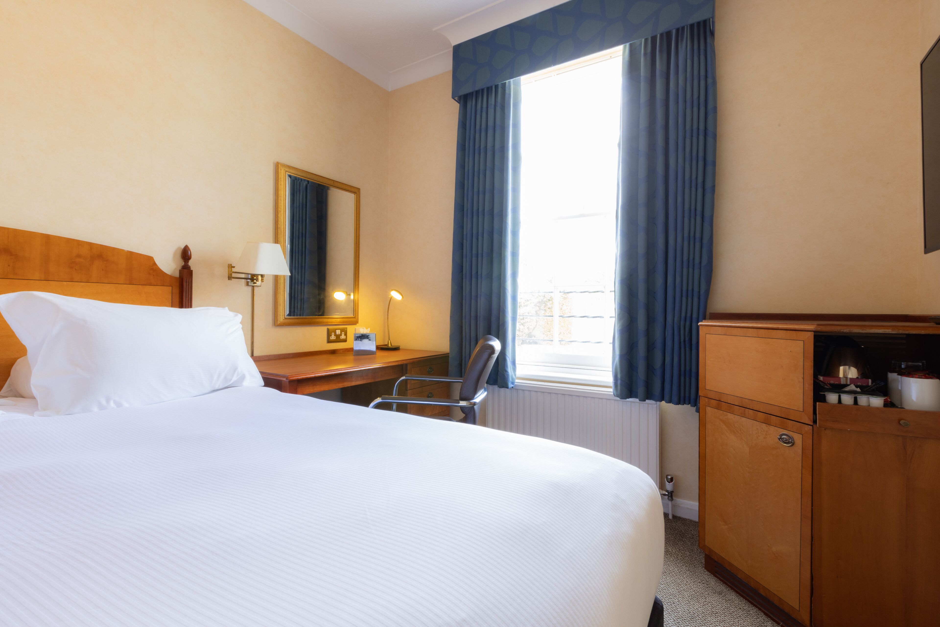 Une chambre d’hôtel avec un grand lit, un bureau avec une lampe, une chaise, une armoire et une fenêtre avec des rideaux.