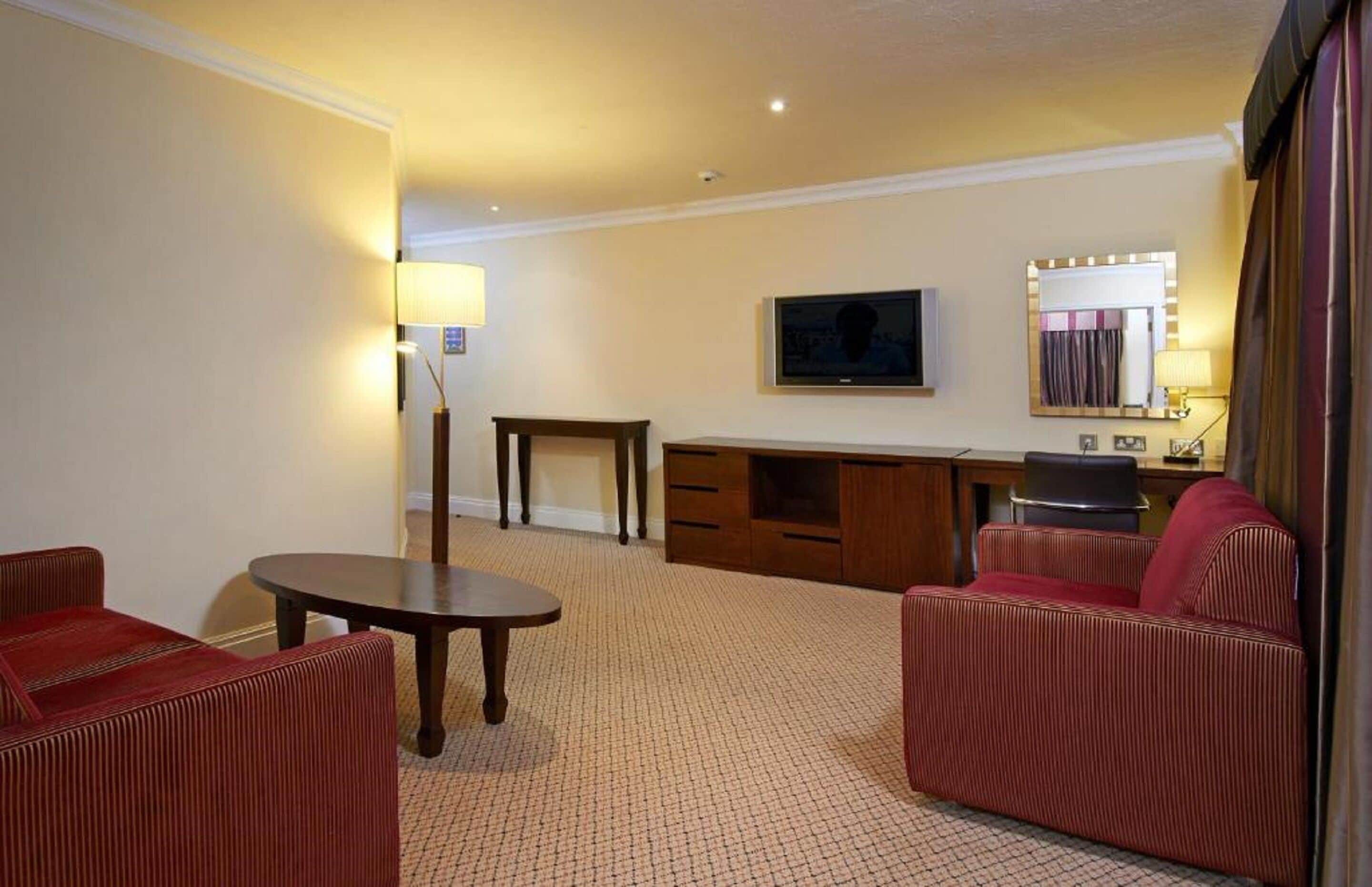 Une chambre d’hôtel avec un canapé, une petite table ronde, une télévision, un miroir et un bureau avec des tiroirs.