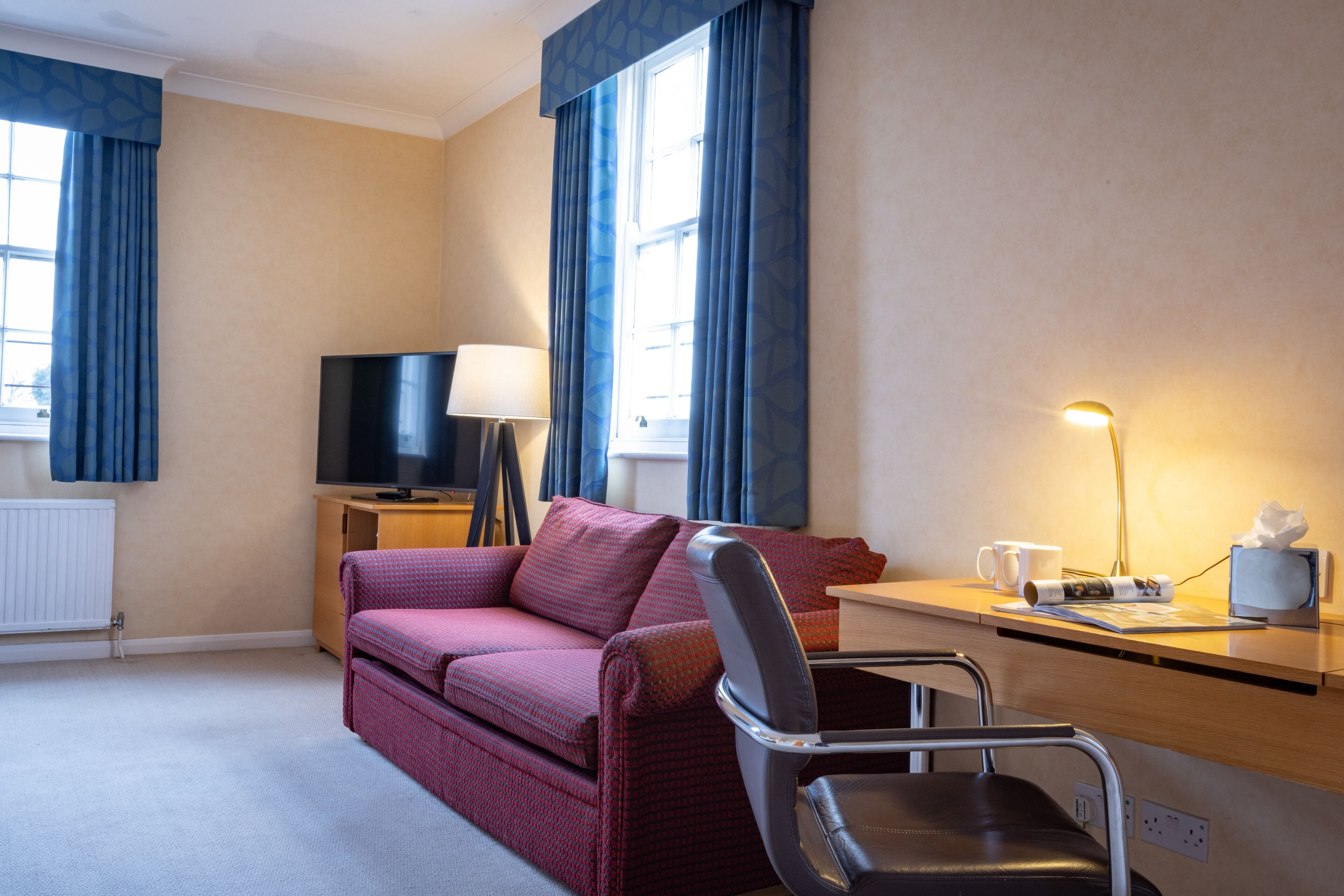 Habitación de hotel con sofá, escritorio, silla, televisor, lámpara y ventana con cortinas.
