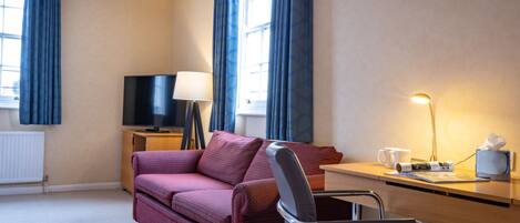 Habitación de hotel con sofá, escritorio, silla, televisor, lámpara y ventana con cortinas.