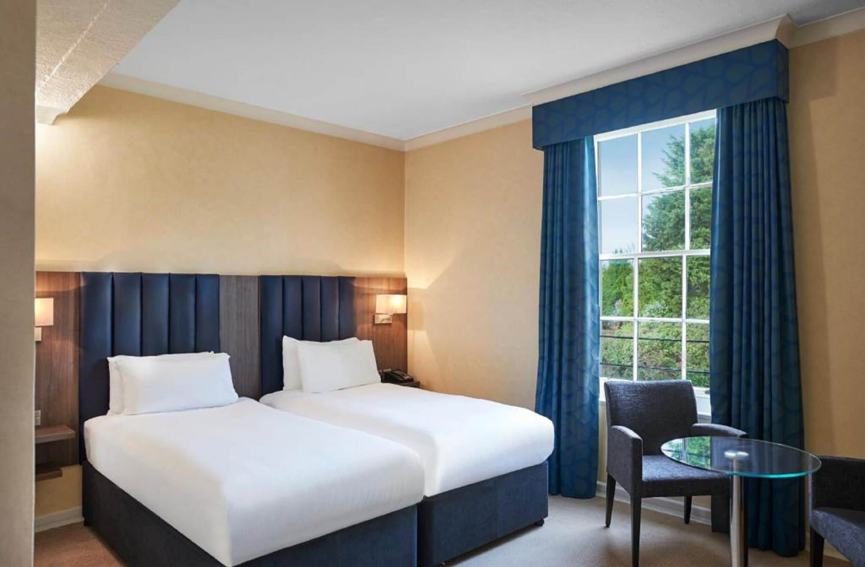 Une chambre d’hôtel avec deux lits, une petite table, une chaise et une fenêtre avec des rideaux.