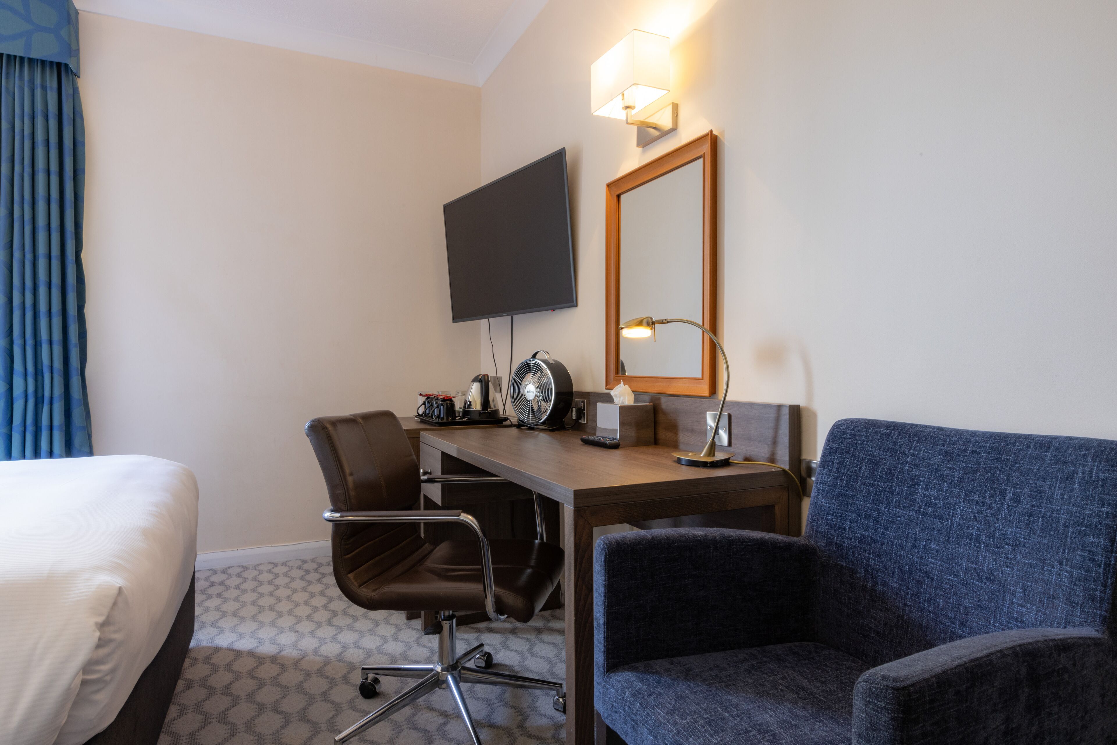 A hotel room with a bed, a desk with a chair, a television, a mirror, and a lamp.