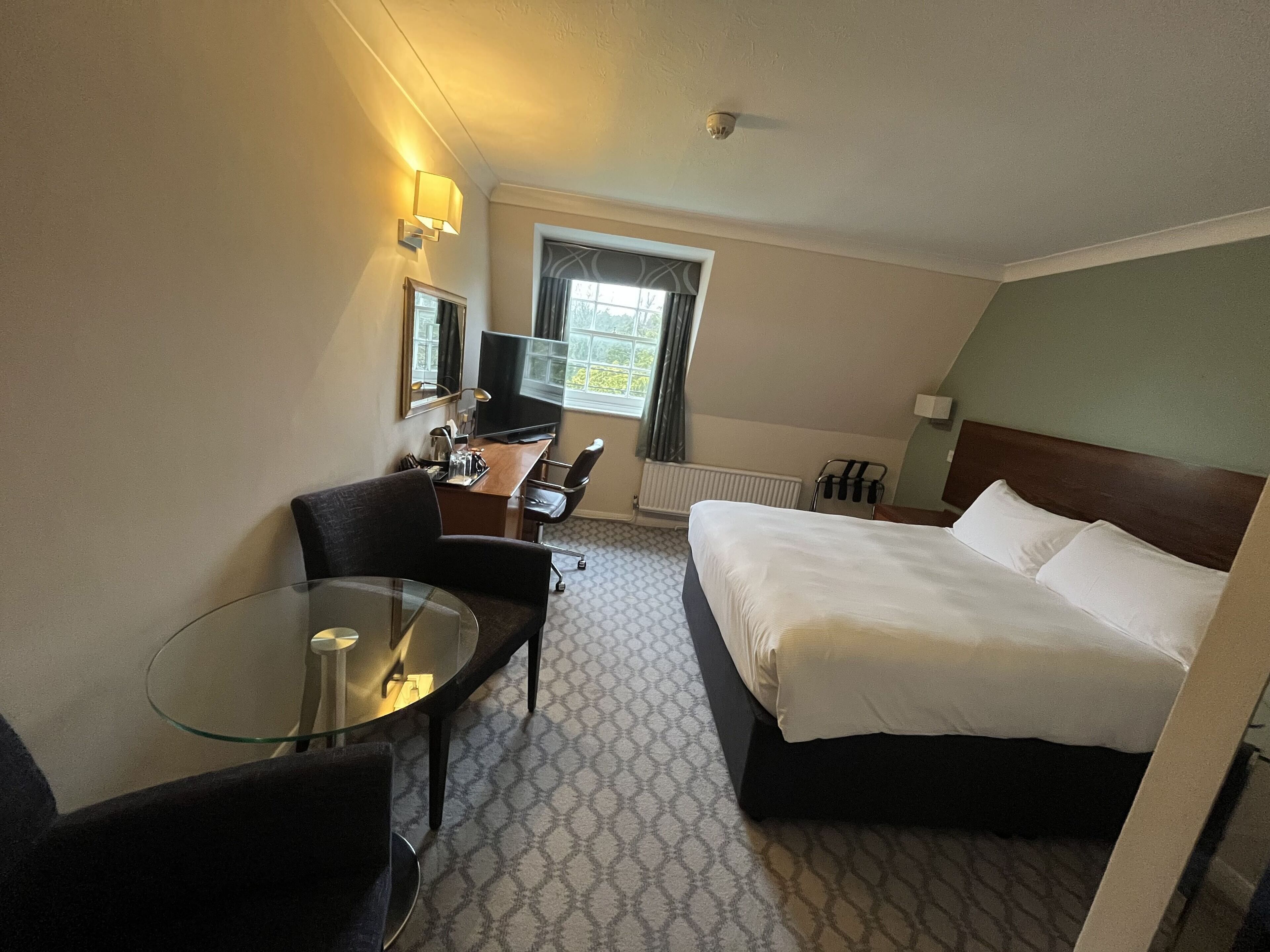 Habitación de hotel con cama, escritorio con computadora, silla, mesita de vidrio, ventana con cortinas y radiador.