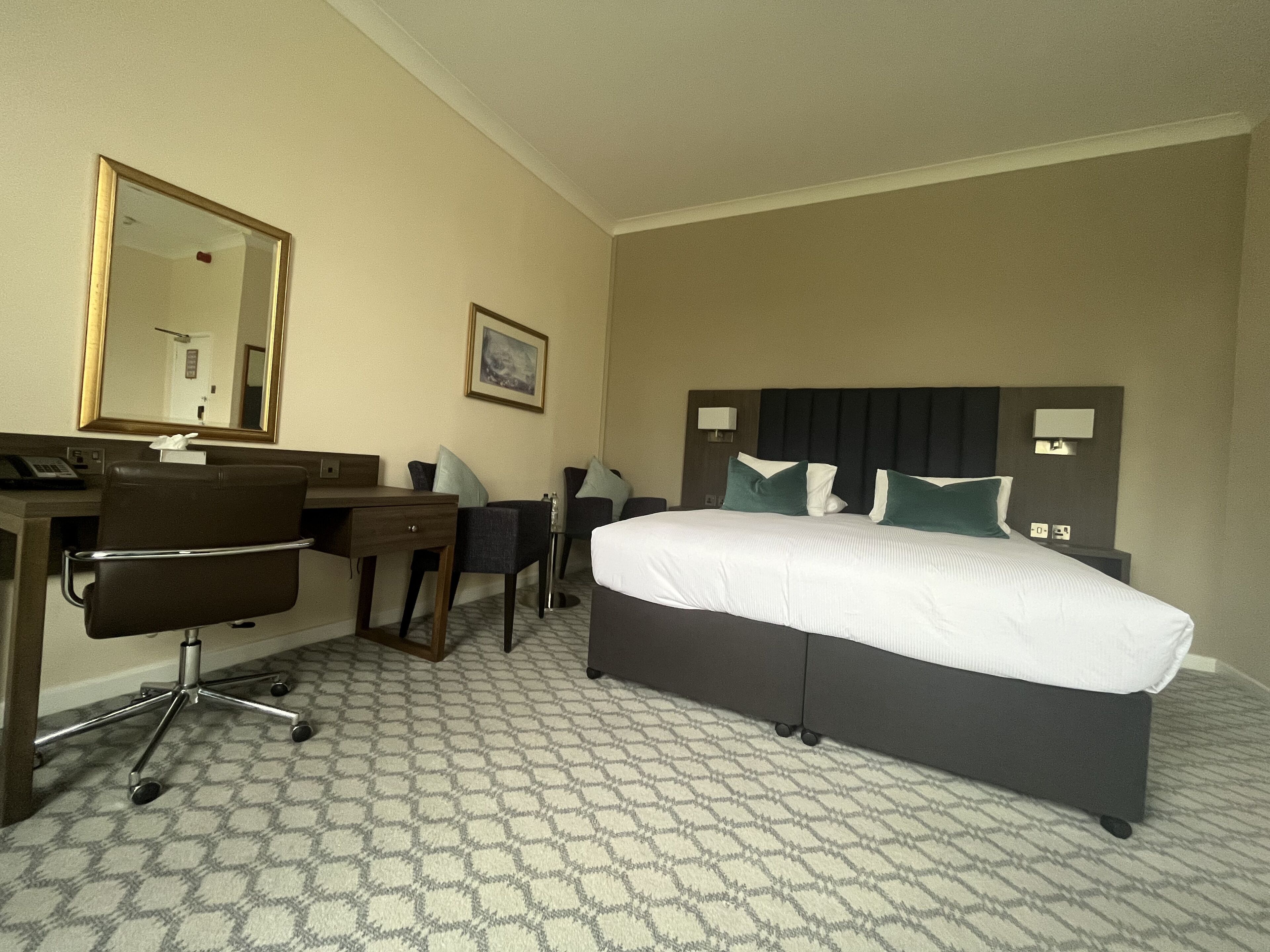 A hotel room with a large bed, a desk with a chair, a telephone, and a mirror.