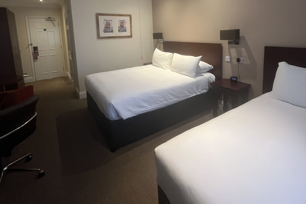 A hotel room with two beds, a desk, a chair, and a door.